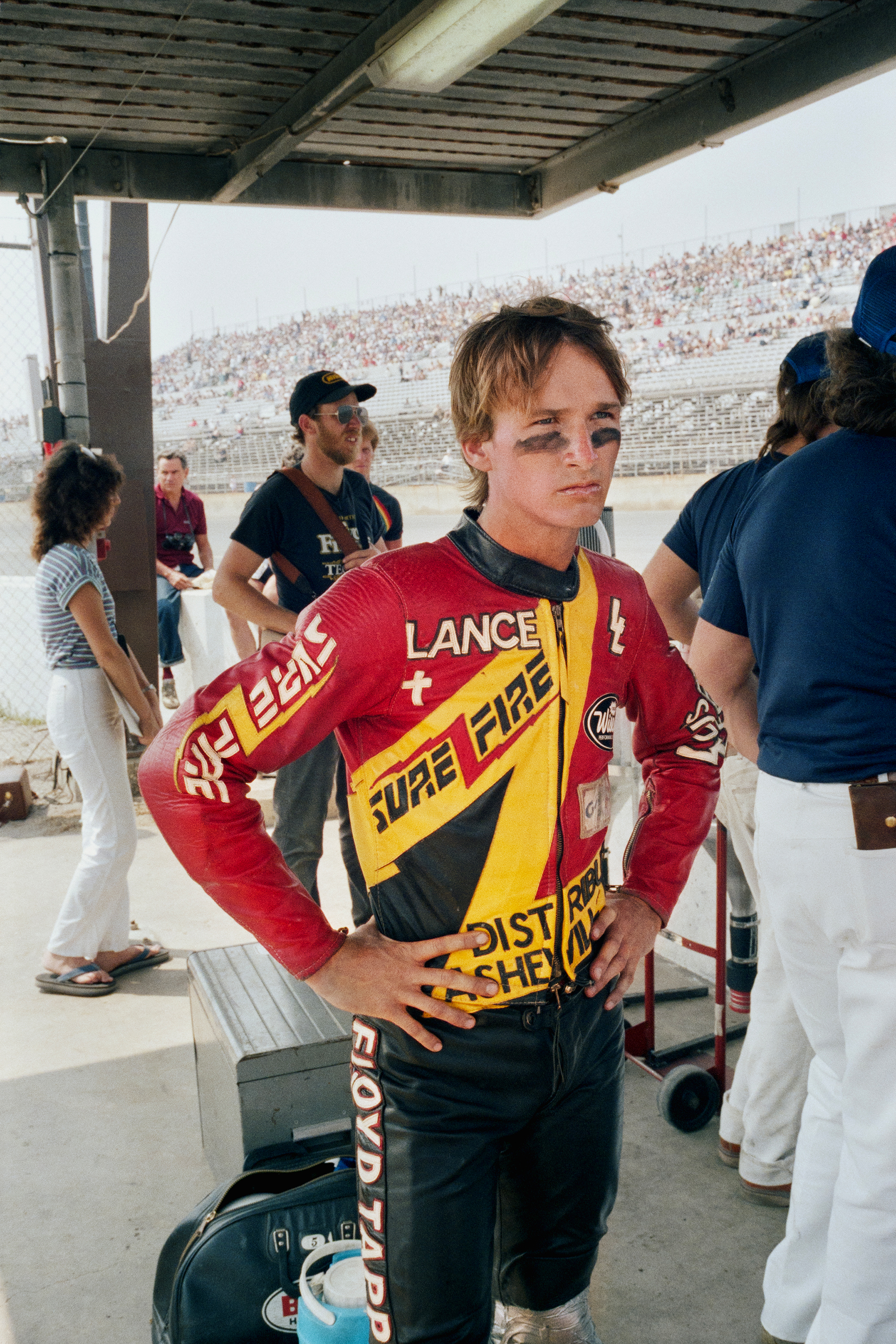 Young motocross rider in red and yellow racing jersey stands under pit tent, stadium crowd visible in background.