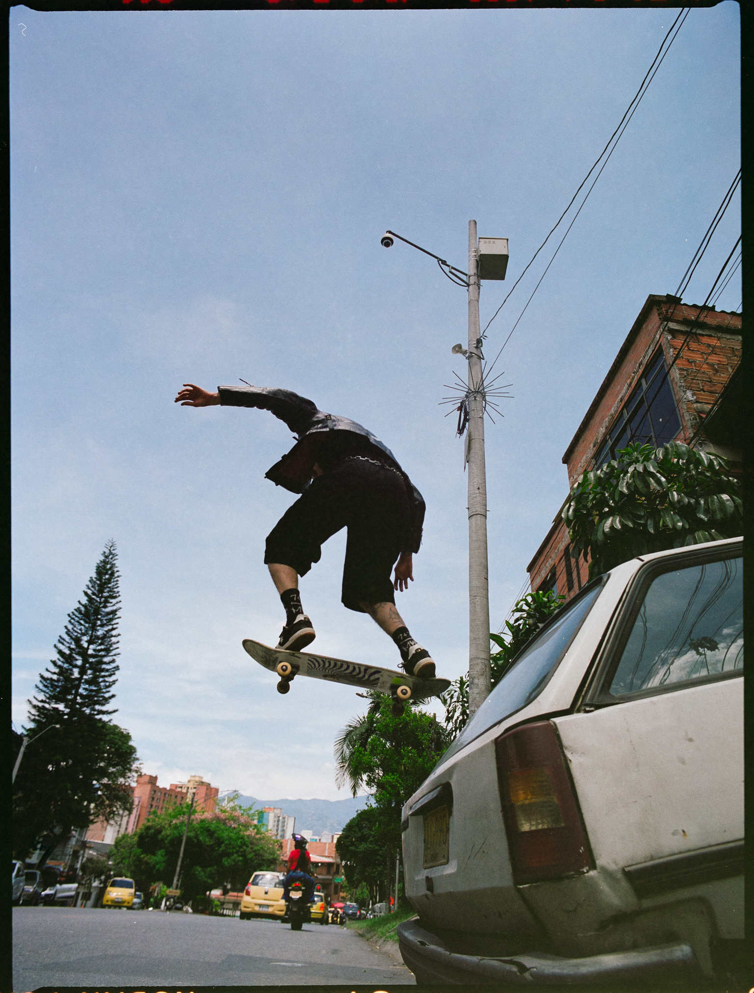 Skateboarder mid-air performing trick on residential street with power lines, palm trees, and parked van visible below blue sky.