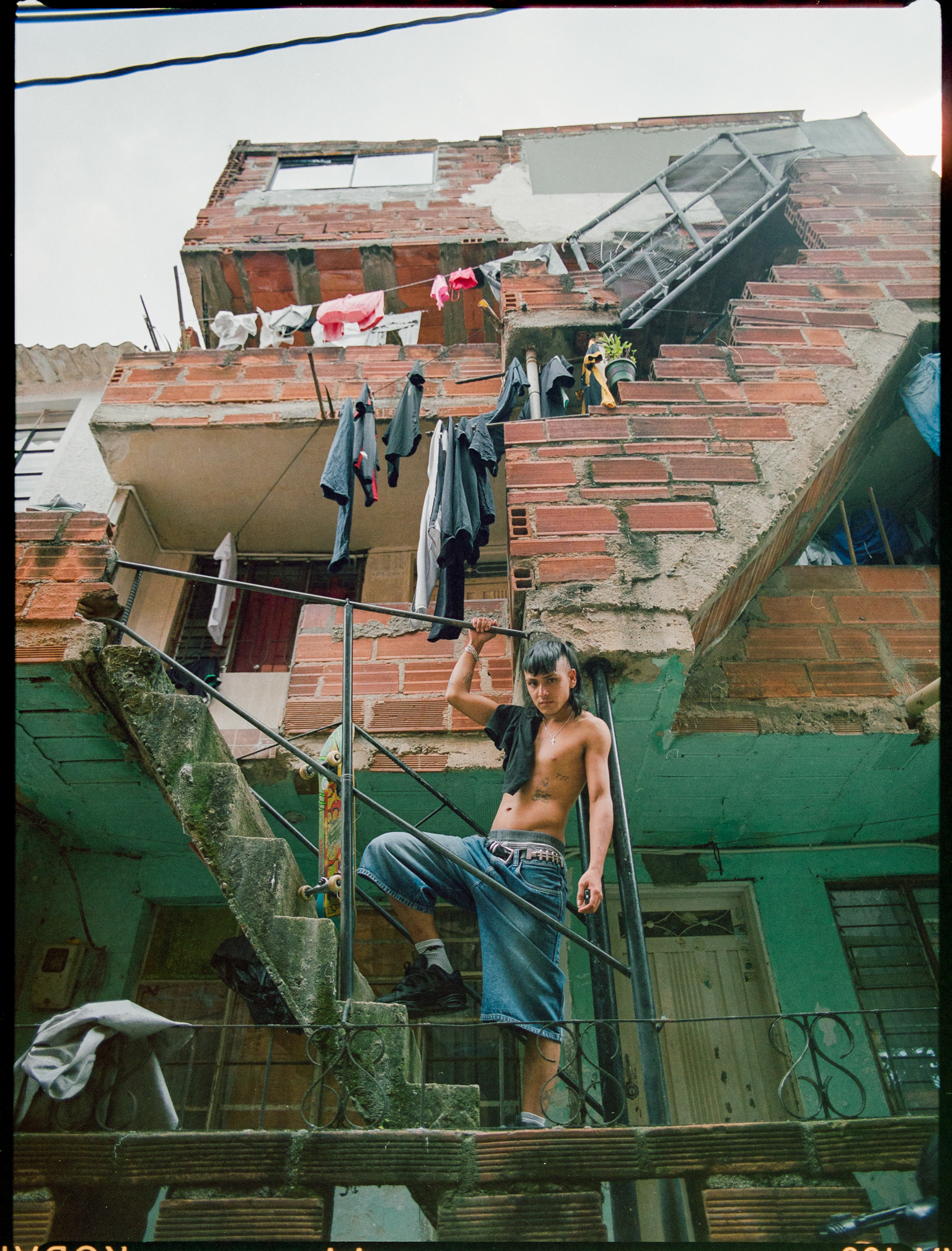 Shirtless man in jeans sits on metal stairs of multi-level brick building with washing lines and makeshift roofing visible.