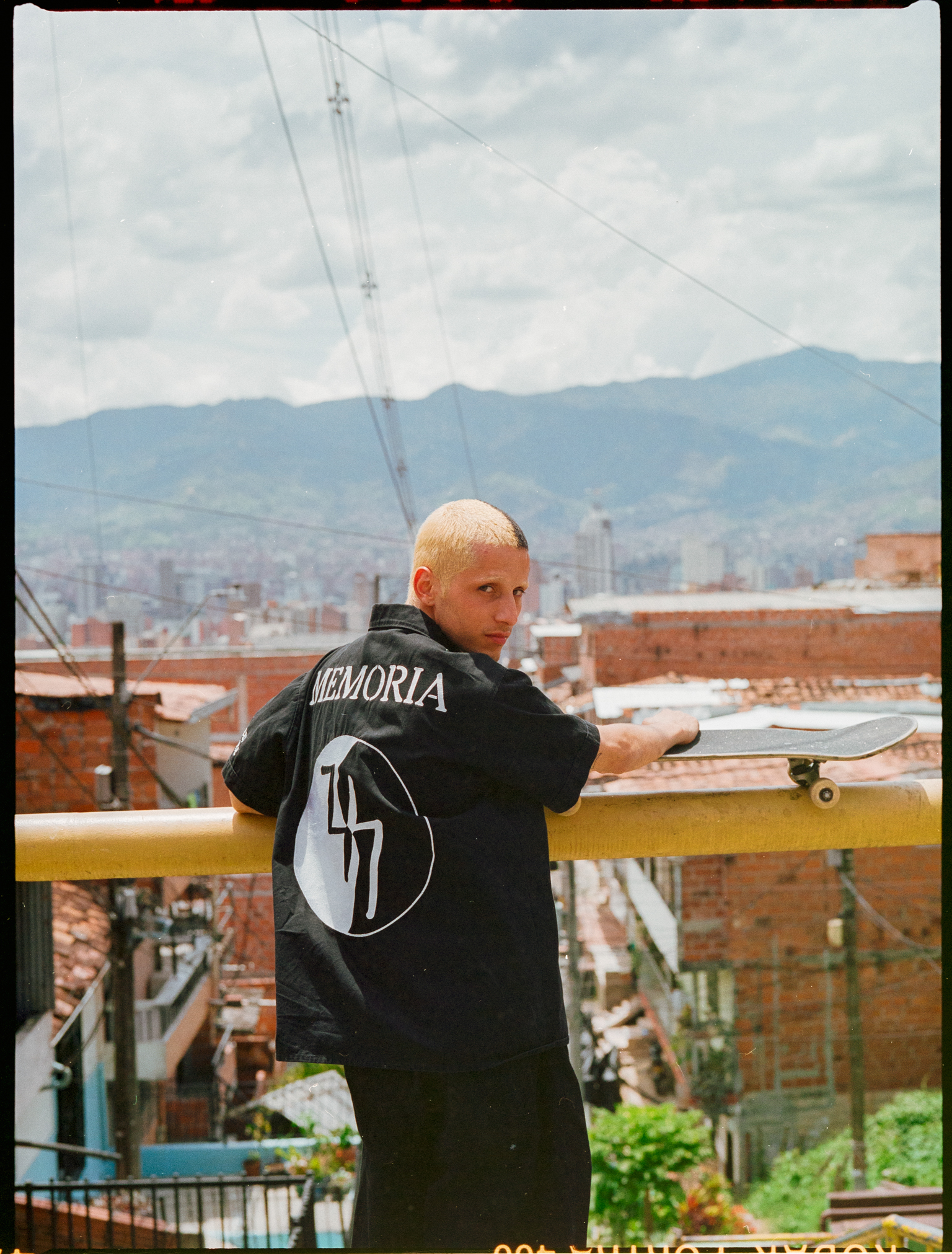 Bald man in black hoodie with "MEMORIA" text and circular logo stands on balcony overlooking city with mountains in background.