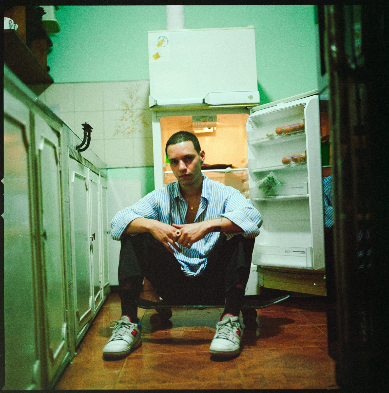 Man in striped shirt sitting on kitchen floor in front of open fridge, wearing white trainers, green-tinted lighting throughout scene.