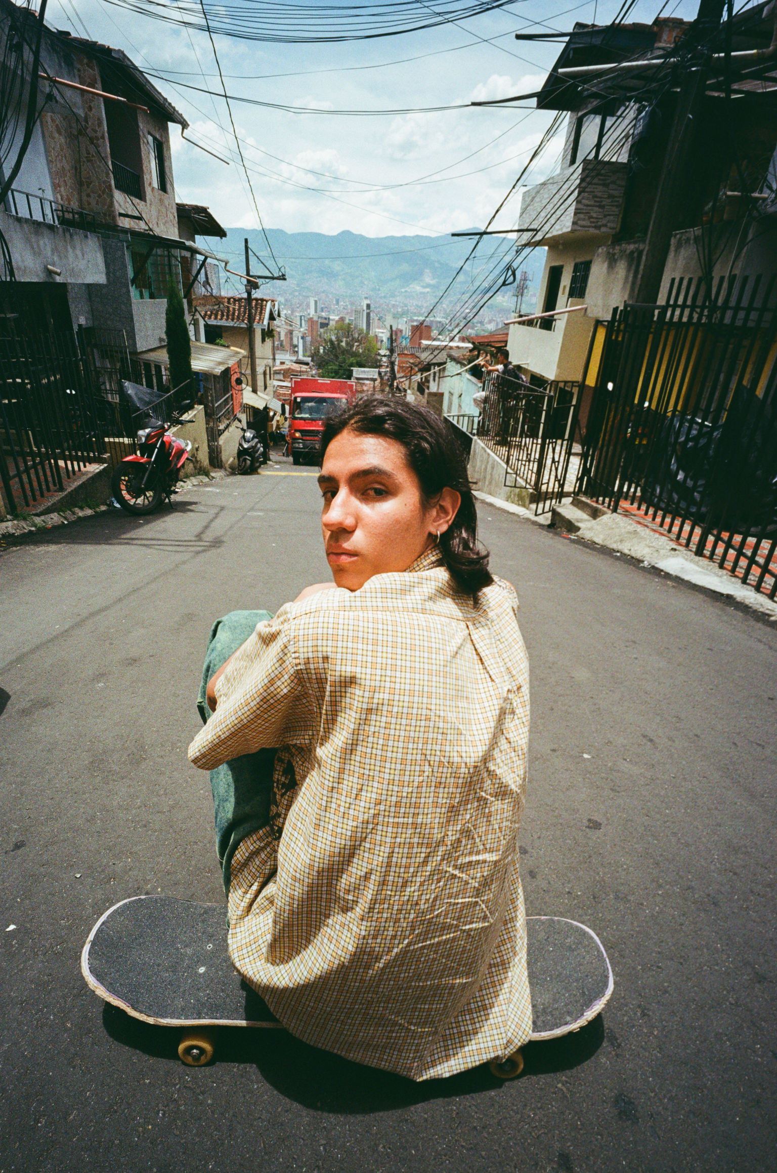 Person in beige checkered shirt sitting on skateboard on steep street with power lines, colourful buildings, and city view below.