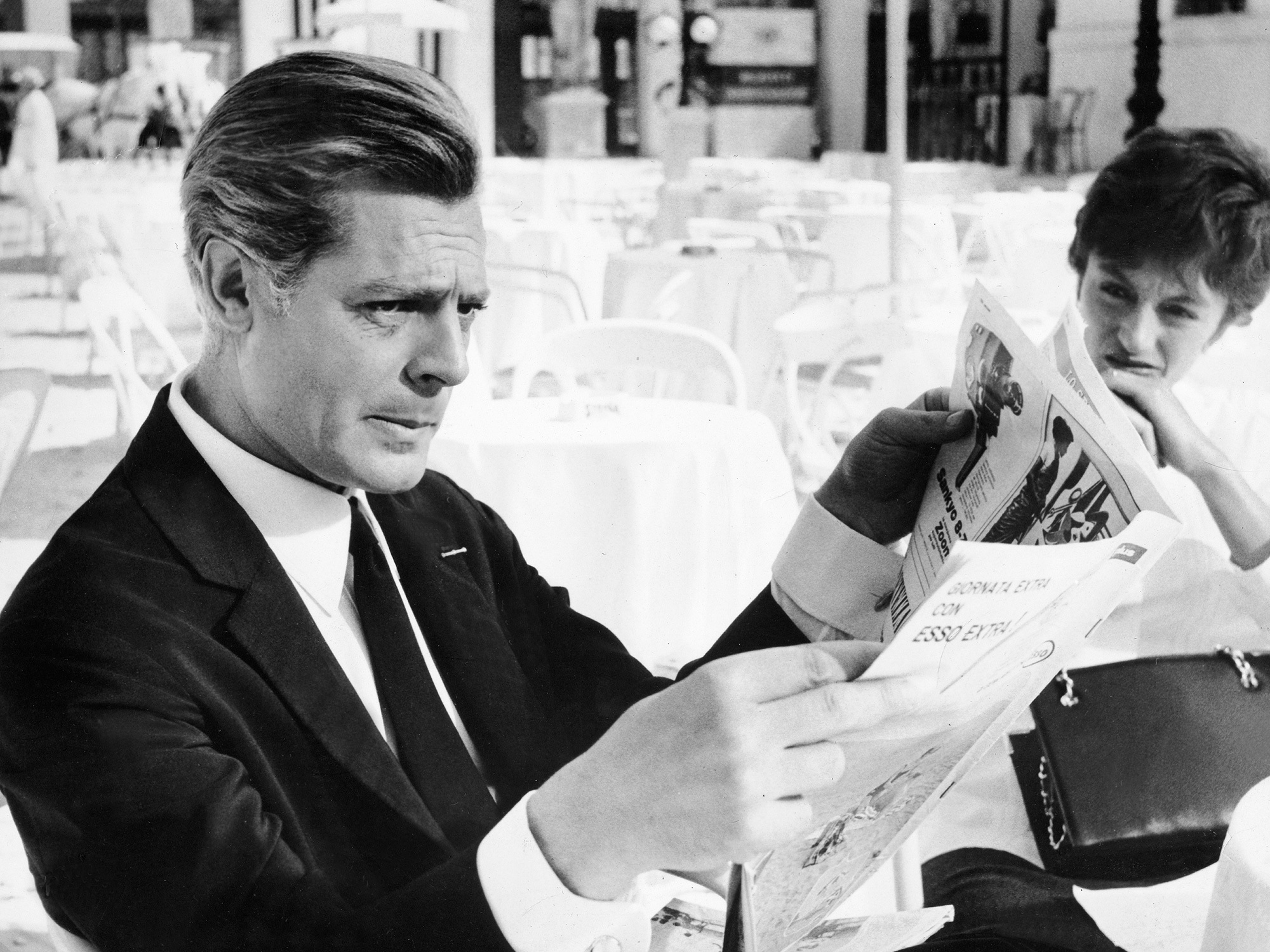 Black and white image of man in dark suit reading newspaper whilst woman reads magazine in background, both seated indoors.