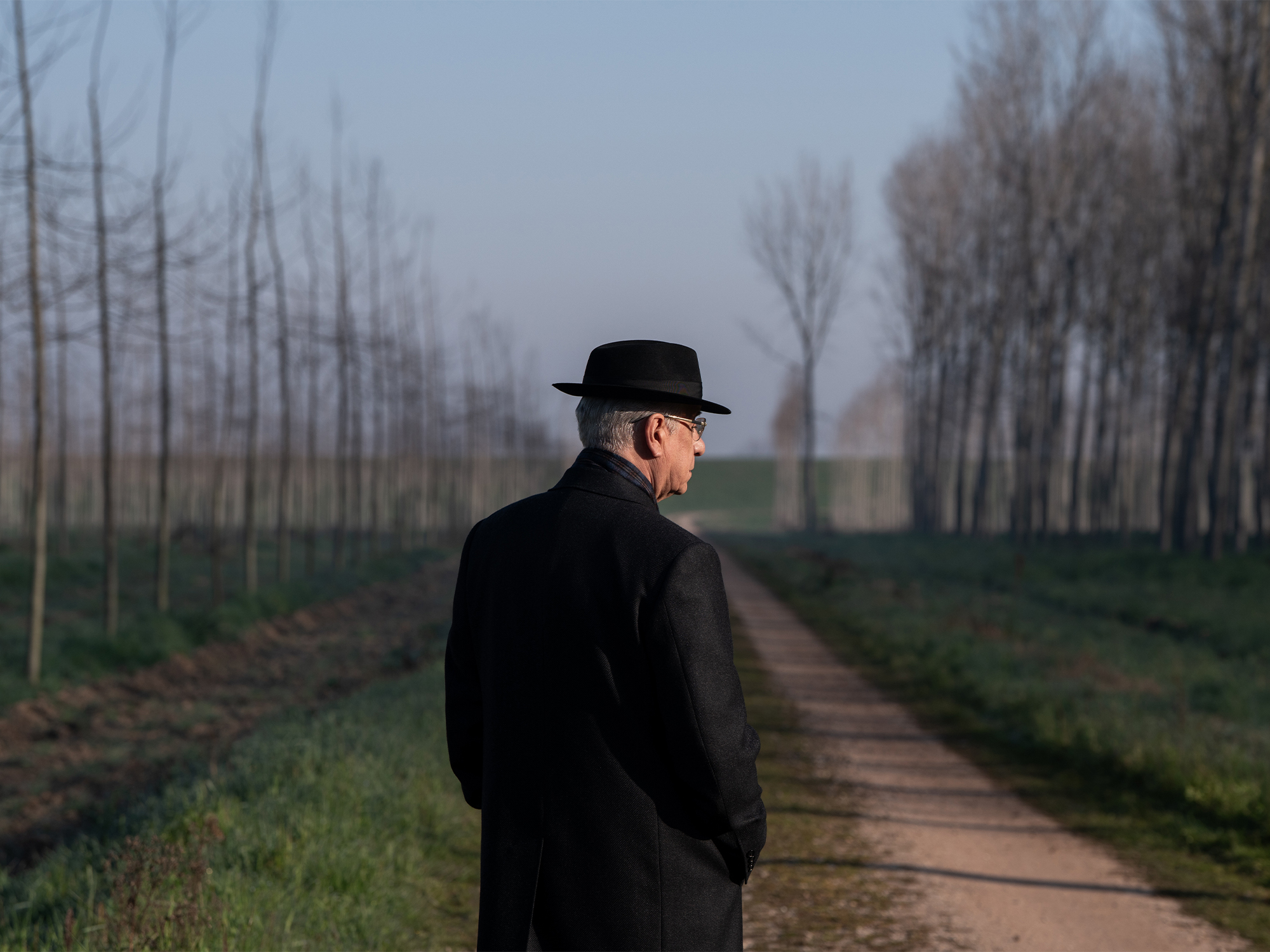 Man in black coat and bowler hat walking on wet path between bare trees, viewed from behind on overcast day.