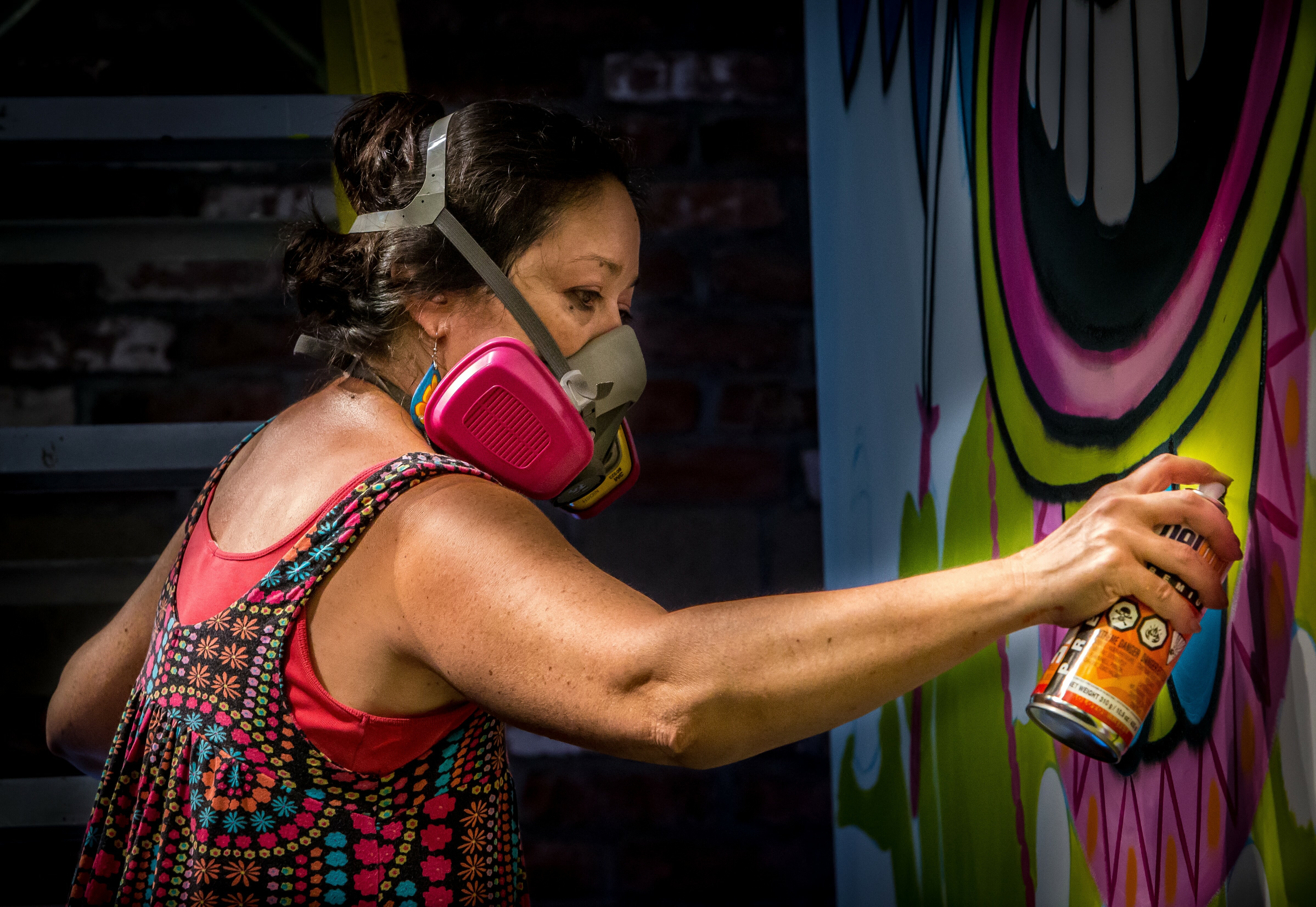 Masked woman in red patterned top spray-painting colourful mural with blue, pink, yellow, and green curved shapes on wall.