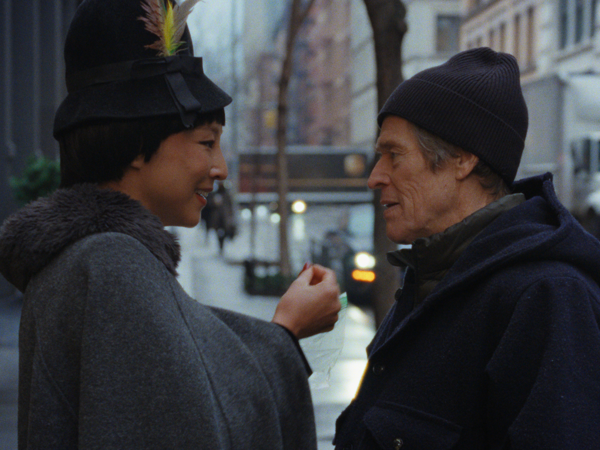 Two people in winter hats and coats face each other on snowy city street with blurred buildings and warm lights in background.