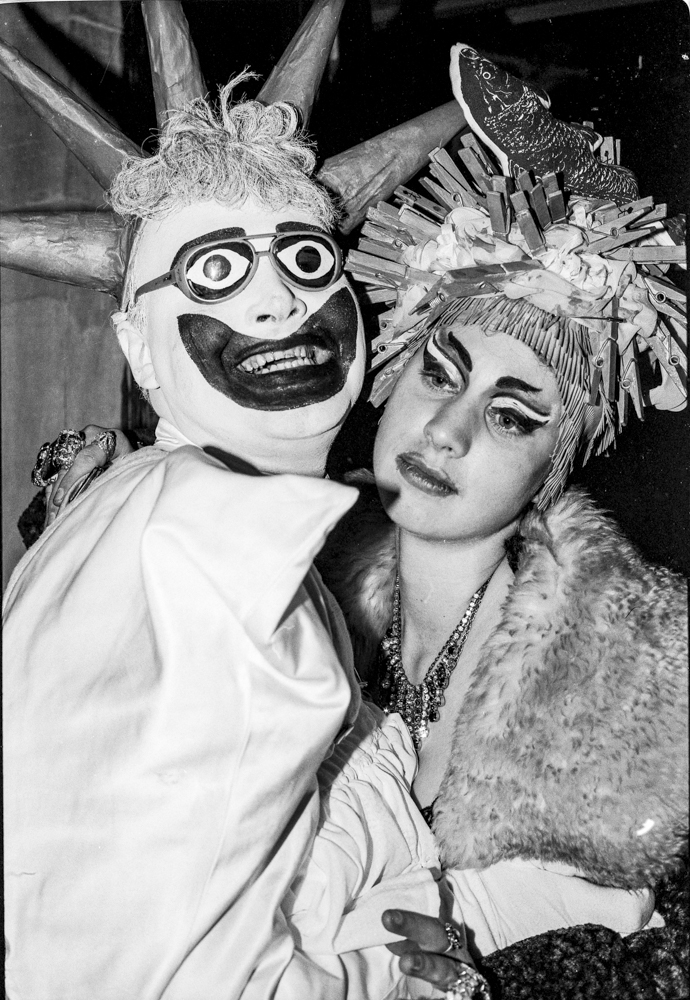 Two people in fancy dress costumes at party - one wearing theatrical mask with large eyes, other in feathered headdress, black and white.
