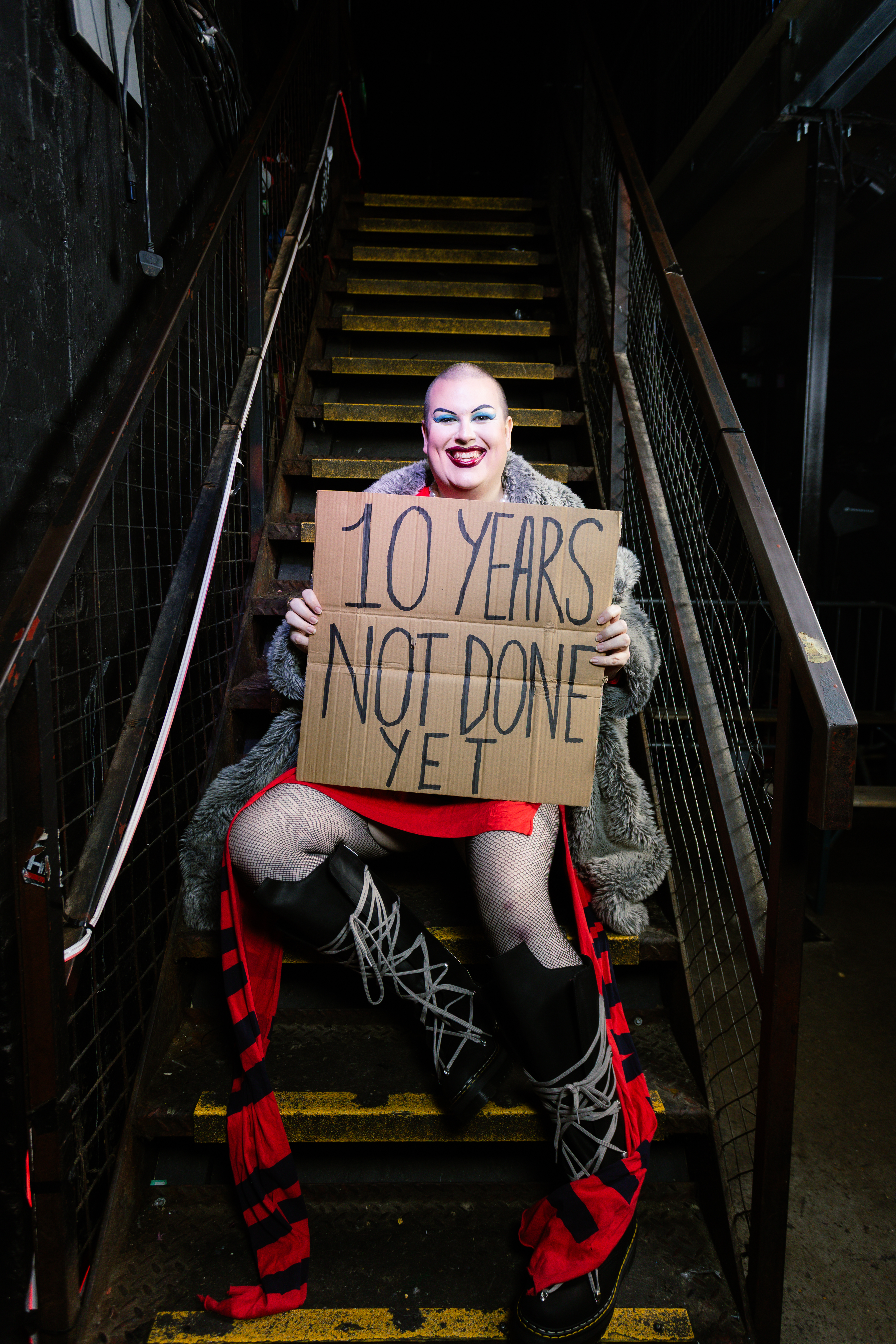 Person in theatrical makeup and costume sits on wooden stairs holding cardboard sign reading "10 YEARS NOT DONE YET"