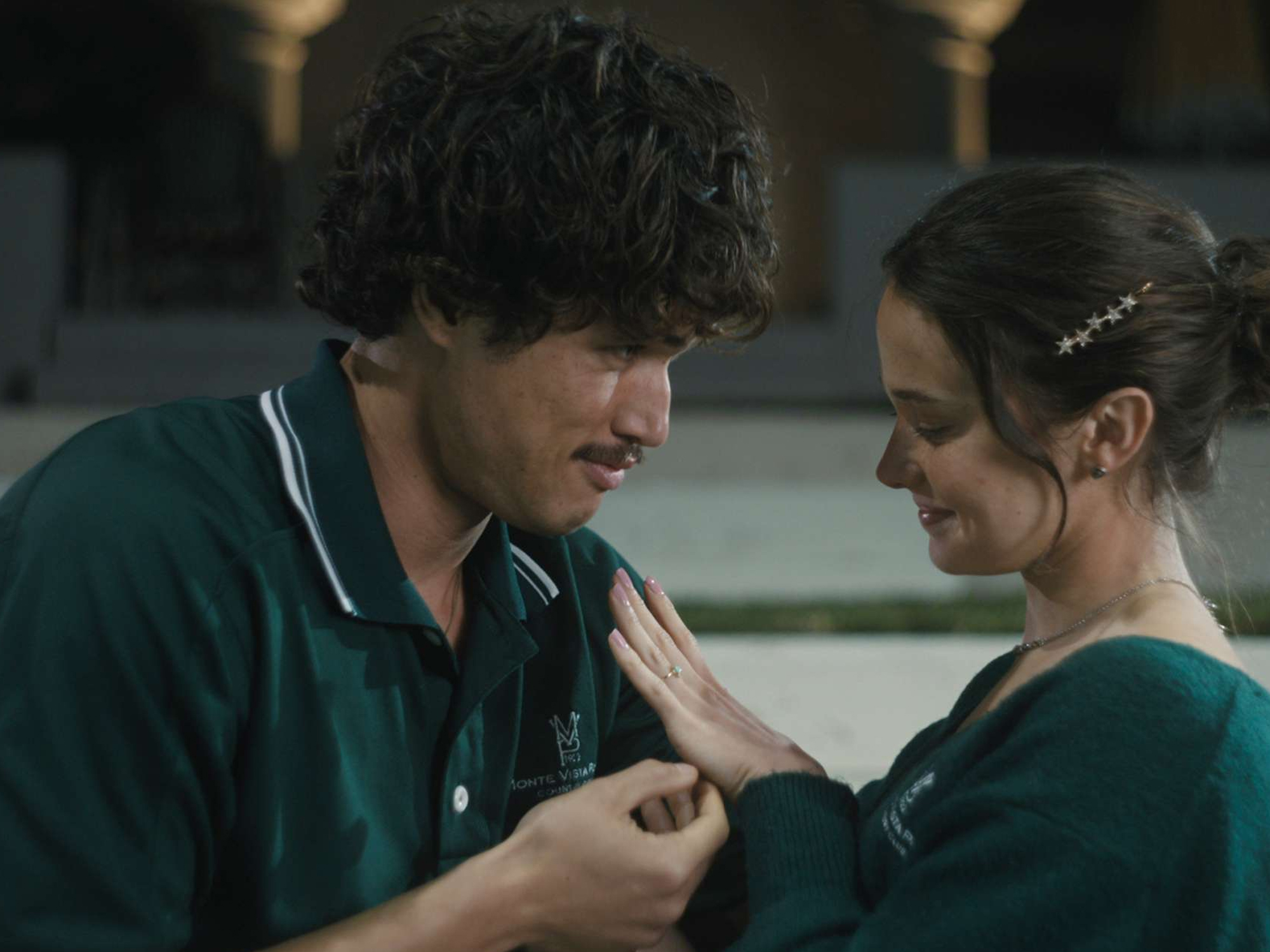 Image depicts a Korean-American man with brown eyes, black hair and a moustache and a white woman with brown eyes and brown hair embracing in a medium shot. The man is wearing a dark green polo shirt and the woman is wearing a dark green jumper. The woman is admiring an engagement ring on her hand, and both are smiling.