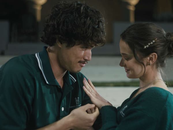 Image depicts a Korean-American man with brown eyes, black hair and a moustache and a white woman with brown eyes and brown hair embracing in a medium shot. The man is wearing a dark green polo shirt and the woman is wearing a dark green jumper. The woman is admiring an engagement ring on her hand, and both are smiling.