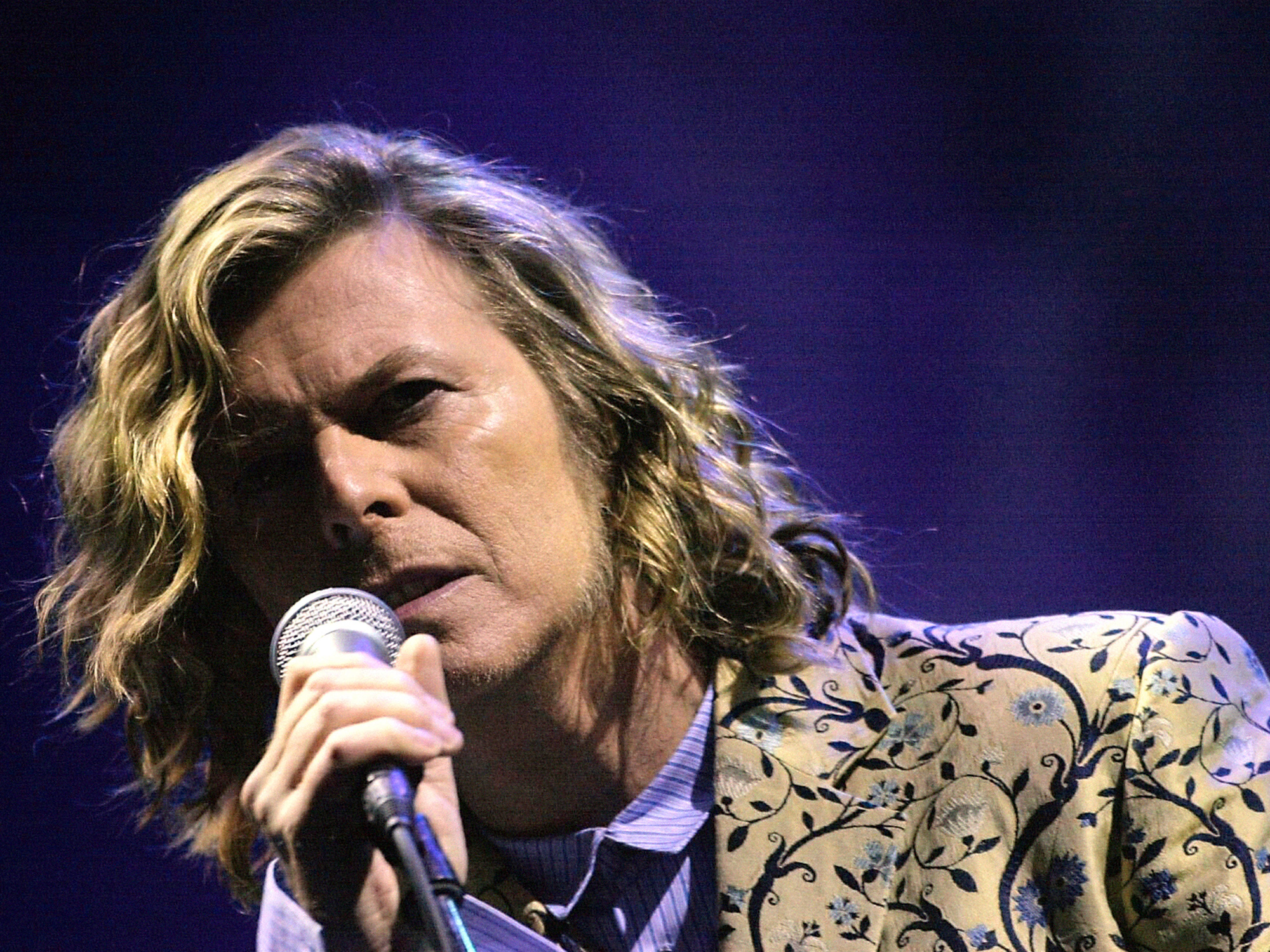 Man with shoulder-length wavy blonde hair singing into microphone, wearing patterned jacket against purple-blue stage lighting.