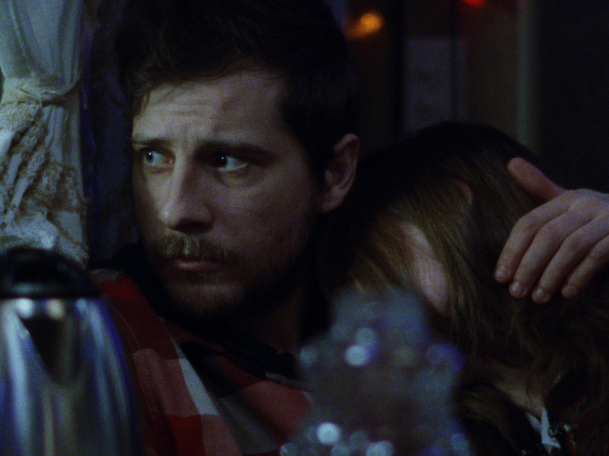 Bearded man in dark clothing embracing someone in dimly lit interior with blurred coloured lights in background.