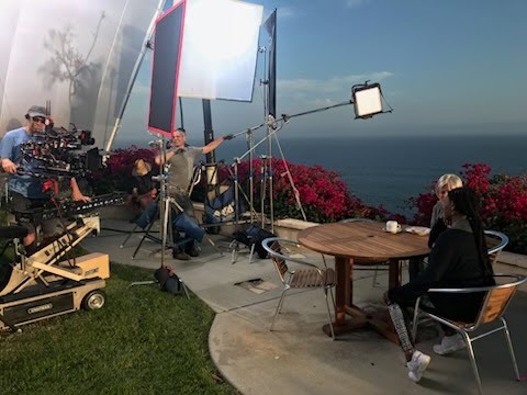 A photograph showing behind the scenes on Grey's Anatomy. A large lighting rig and monitors light a scene of a couple sitting at a table in front of an ocean backdrop.