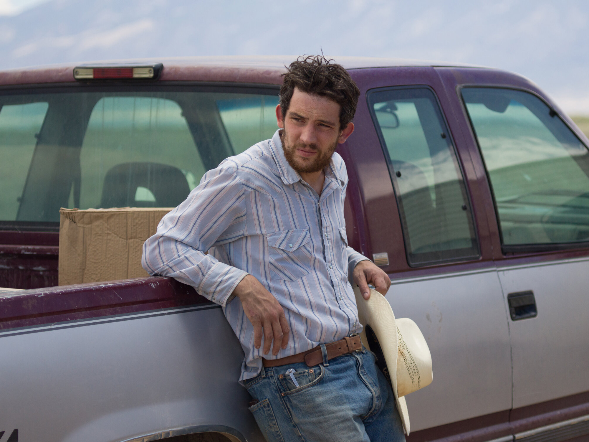 Josh O'Connor stands against a pick-up truck, holding a cream cowboy hat in his hand.