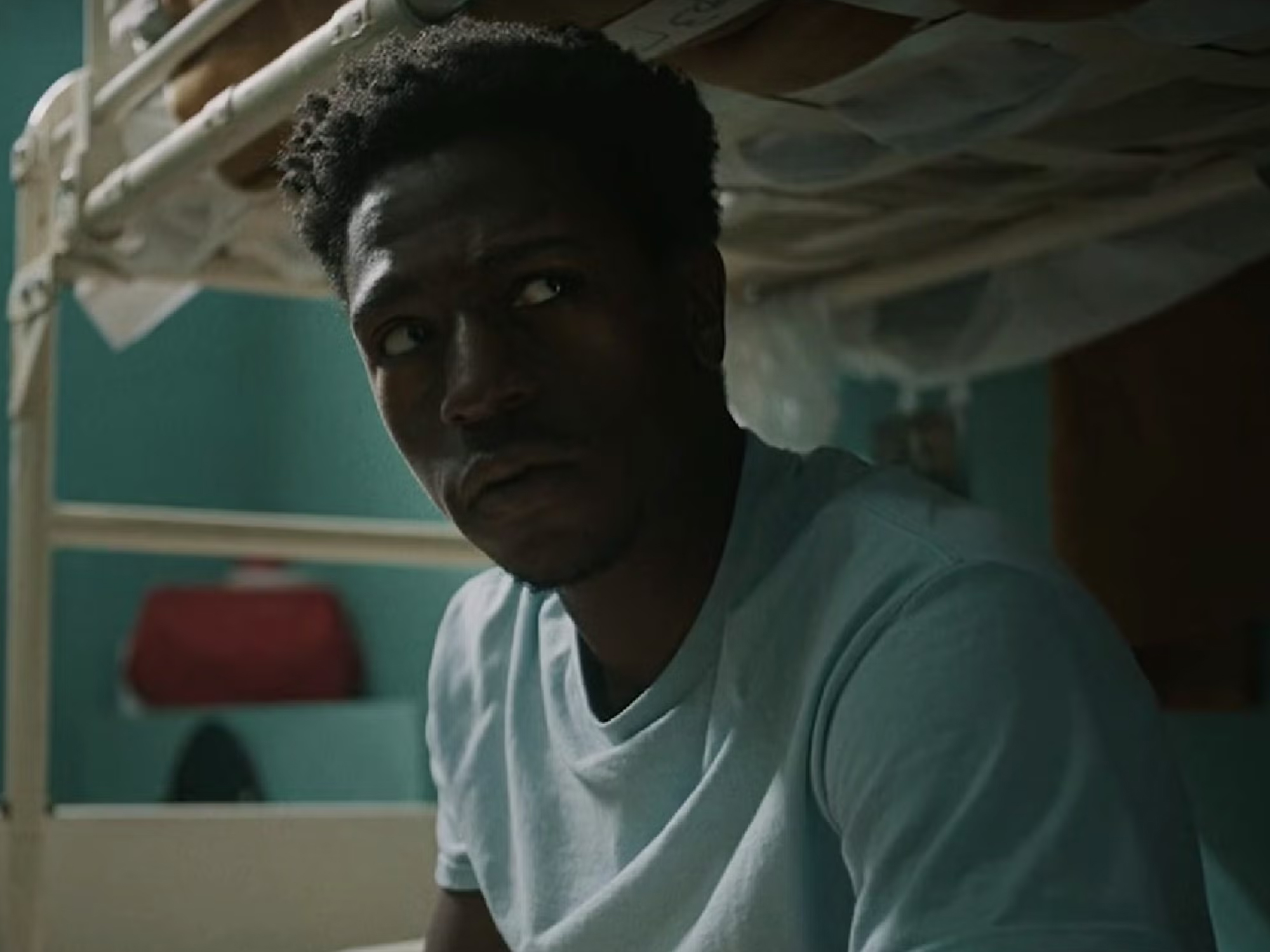David Jonsson, a Black British actor, in the film Wasteman. David wears a grey t-shirt and is sitting on the bottom bunk of a bunkbed in a prison cell.