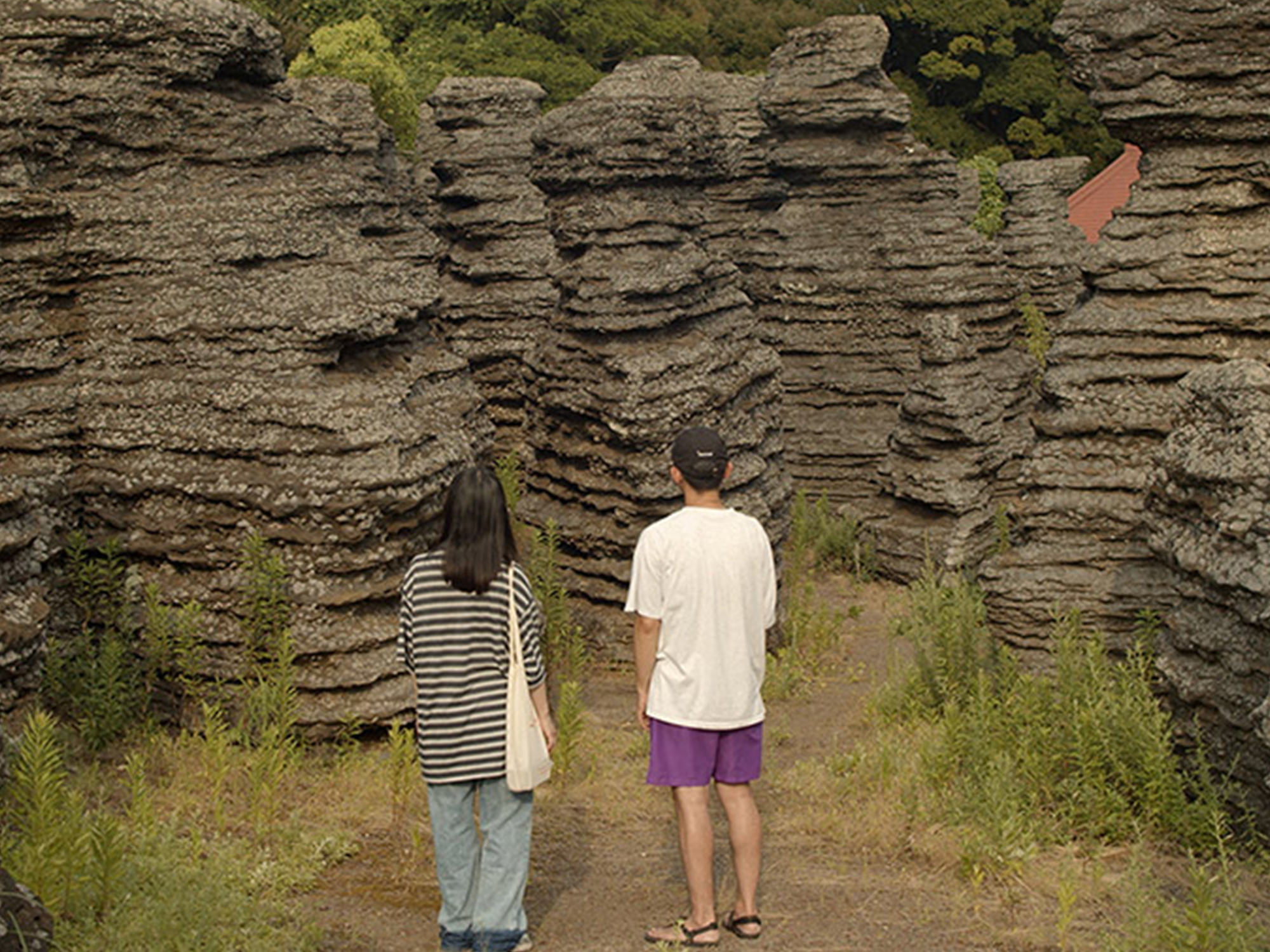 Two people standing on dirt path viewing layered rock cliff face with horizontal sedimentary strata and green vegetation patches.