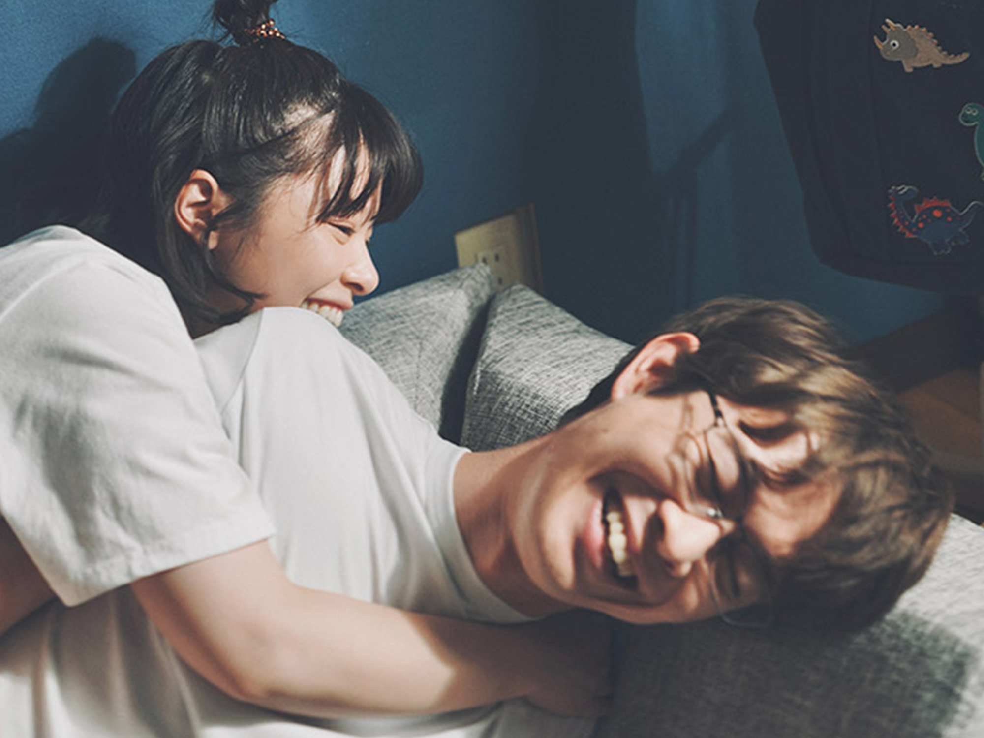 Two people in white shirts lying on a bed, one person laughing whilst covering their face with their hand, the other smiling nearby.