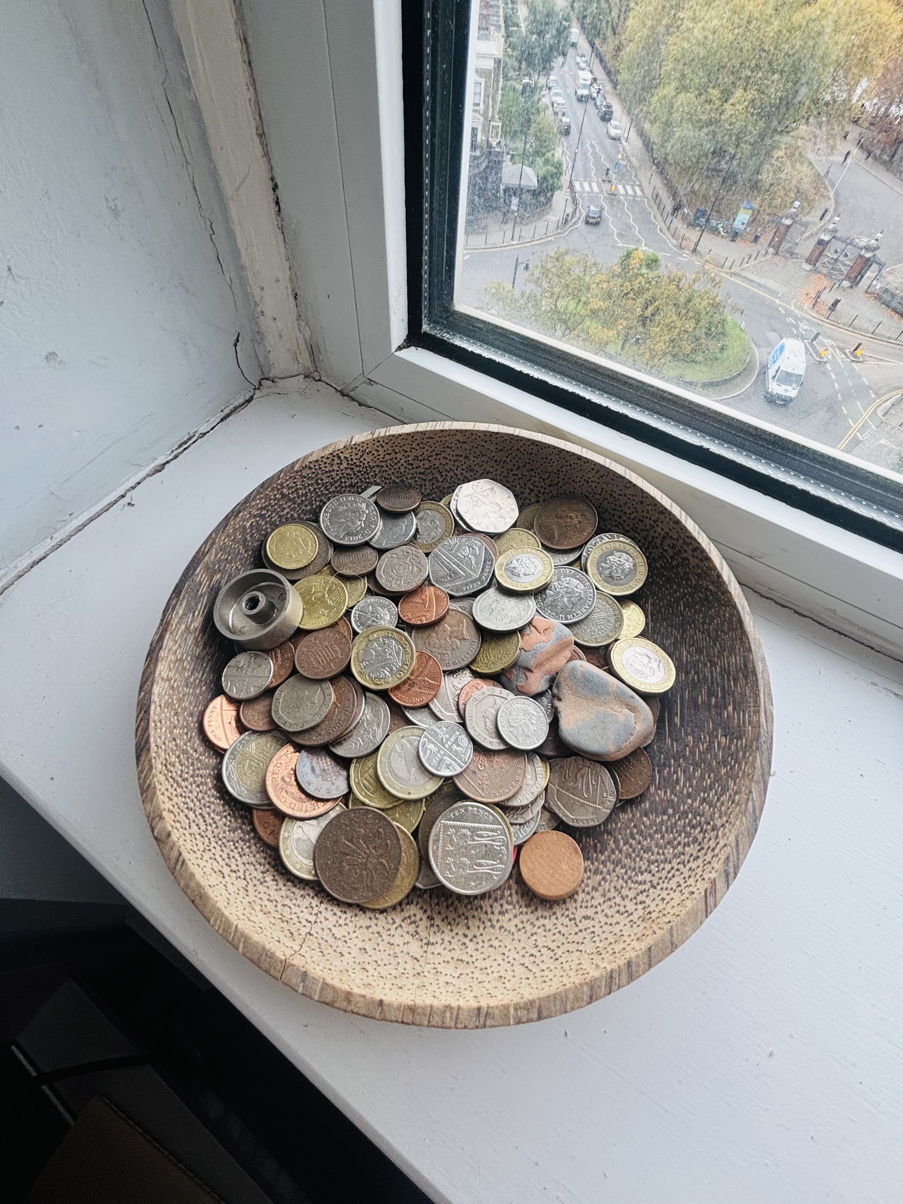 Wooden bowl filled with various coins on windowsill, overlooking street with trees and buildings below.