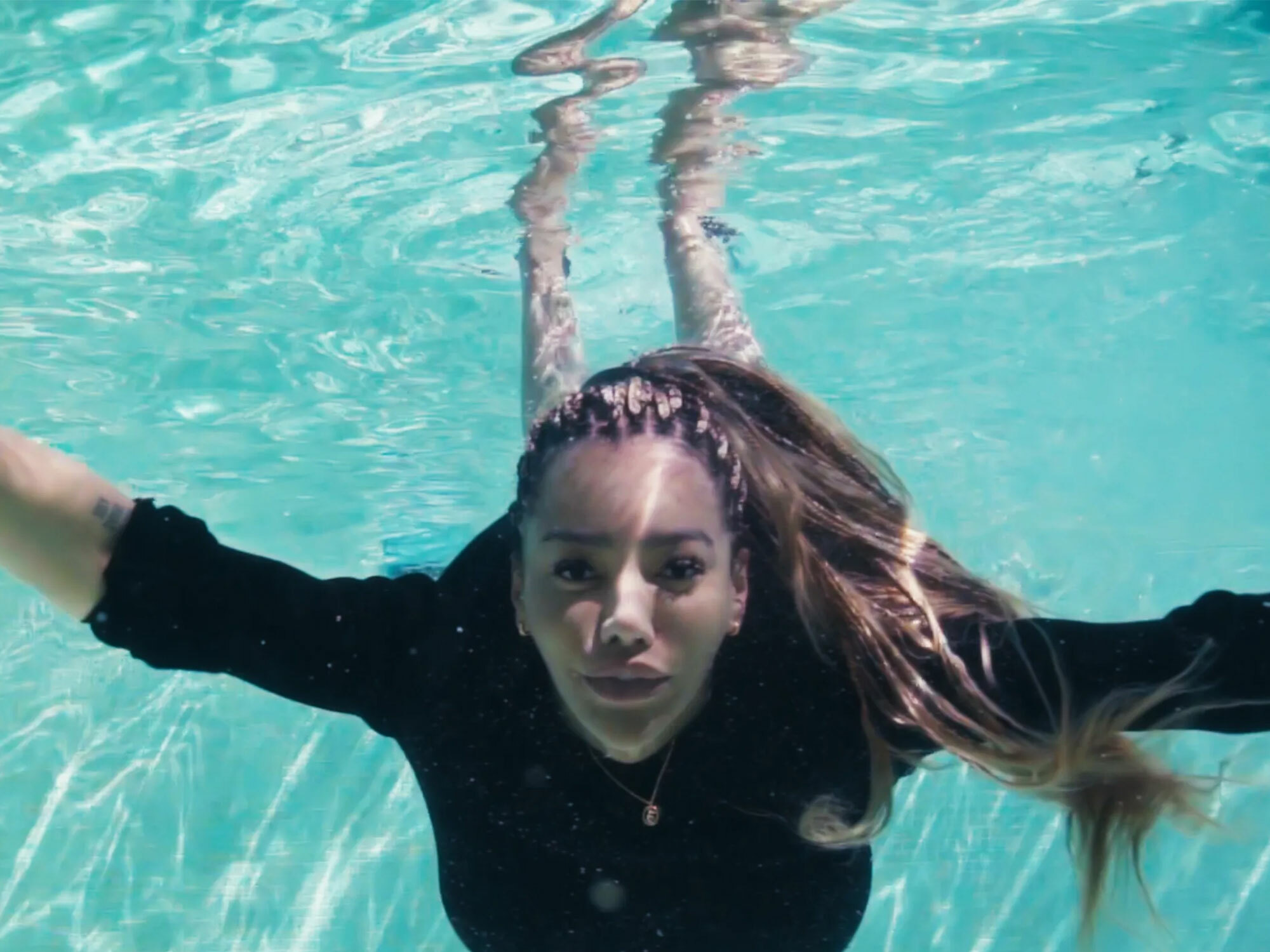 Underwater image of a woman wearing a black wetsuit with her arms outstretched, with long dark hair floating around her in a turquoise-coloured pool.