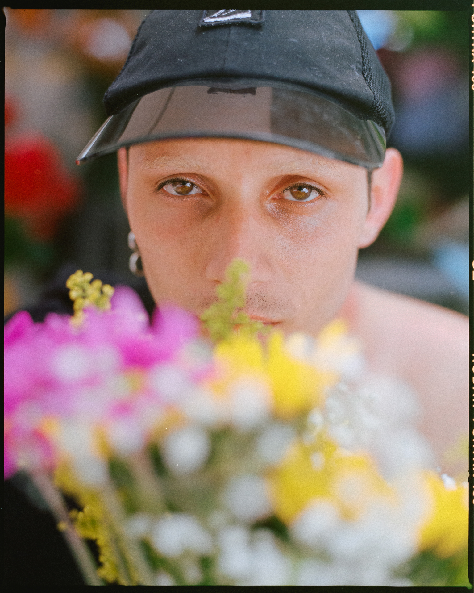 Person in black cap behind blurred pink and yellow flowers in foreground, with soft-focus background.