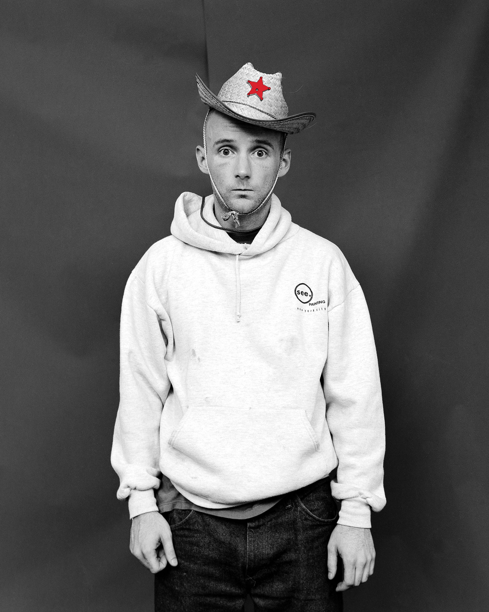 Man in white hoodie and cowboy hat with red star, black and white portrait against grey background.