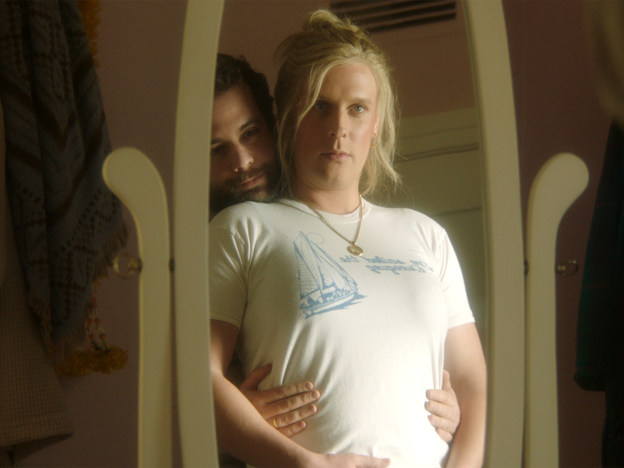 Blonde woman in white t-shirt stands in doorway with person behind her placing hands on her pregnant belly.