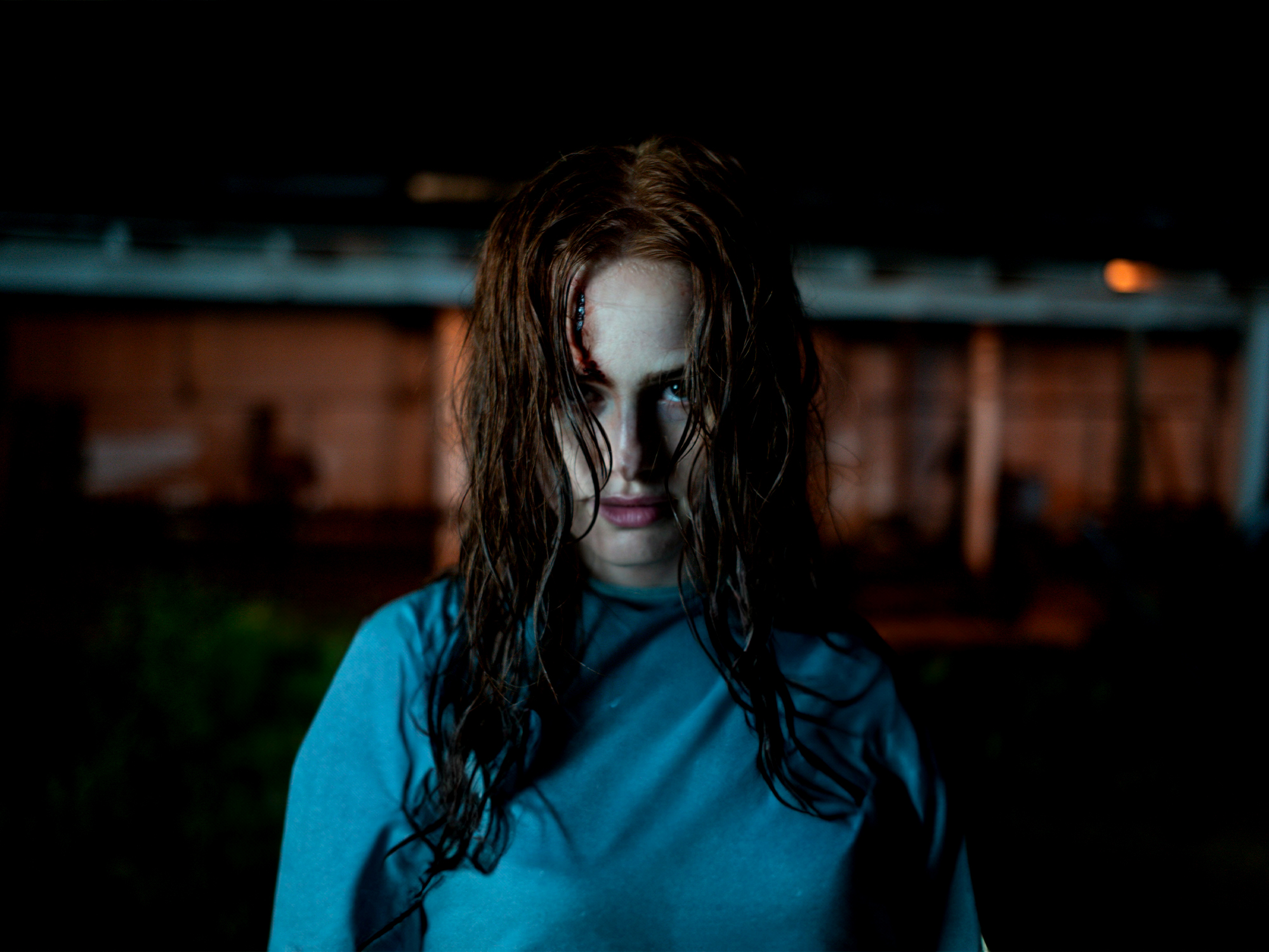 Person with long wet dark hair covering face, wearing blue top, standing outdoors at night with blurred lights behind.