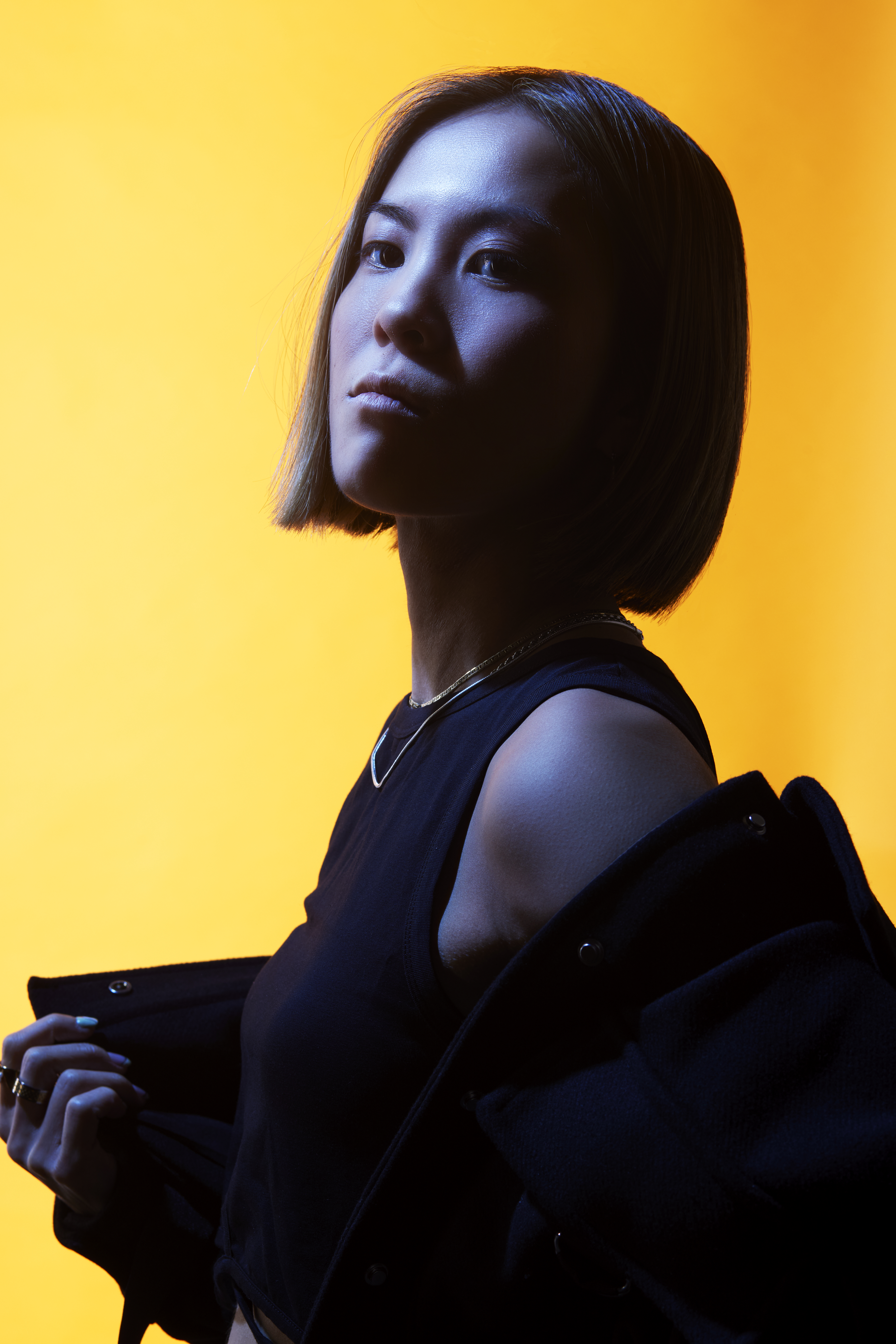 Woman with dark bob haircut in black clothing against bright yellow background, lit with blue and orange tones creating dramatic contrast.