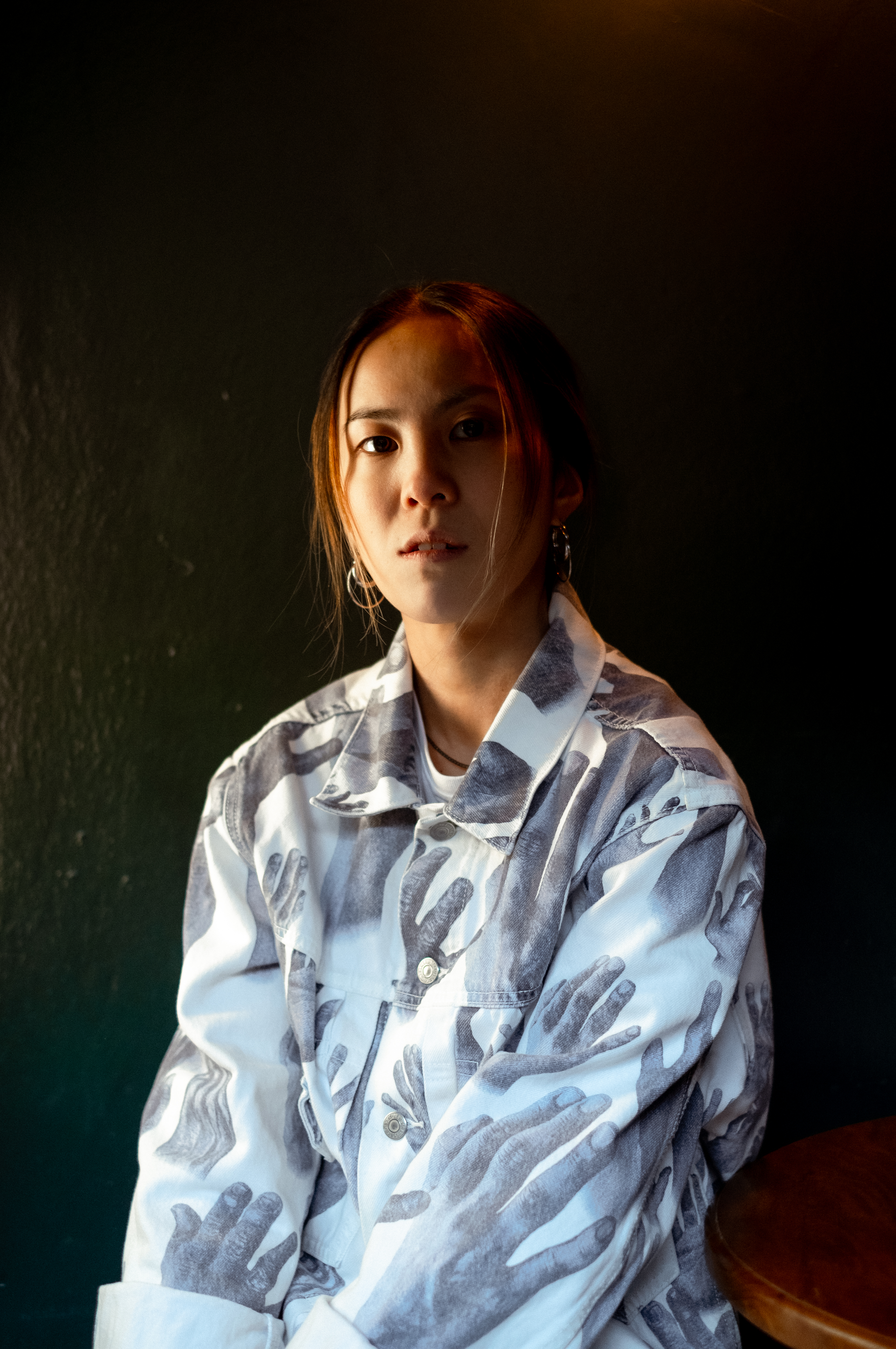 Woman with brown hair wearing oversized white shirt with grey hand print pattern, sitting against dark green wall with warm lighting.