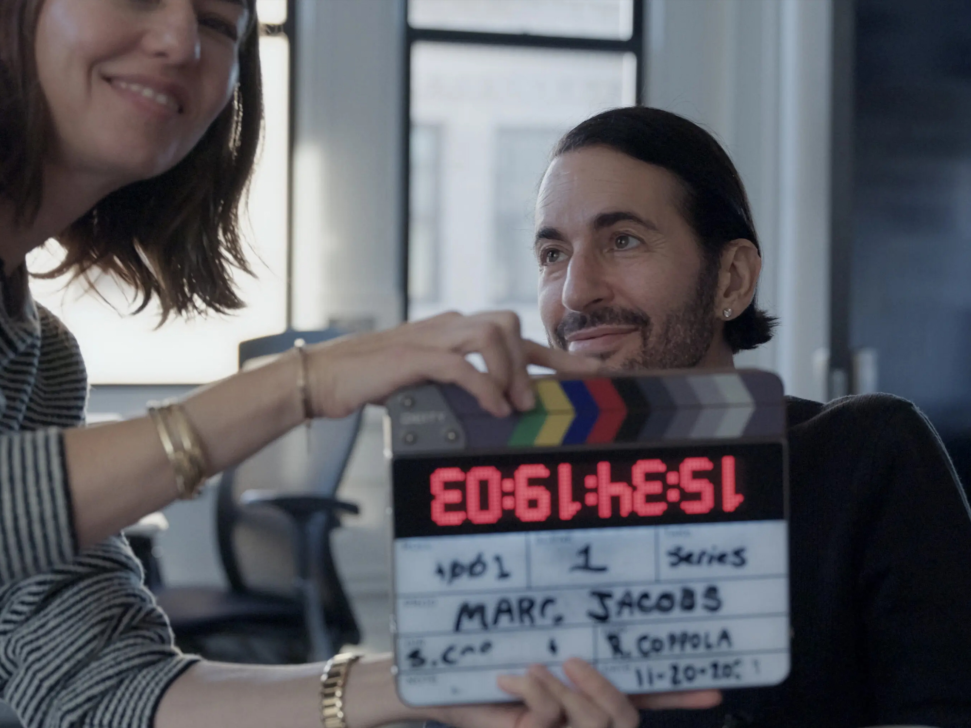 Woman holding film clapperboard reading "E0:61:4E:51" and "MARK JACOBS" whilst man with long dark hair sits behind her.