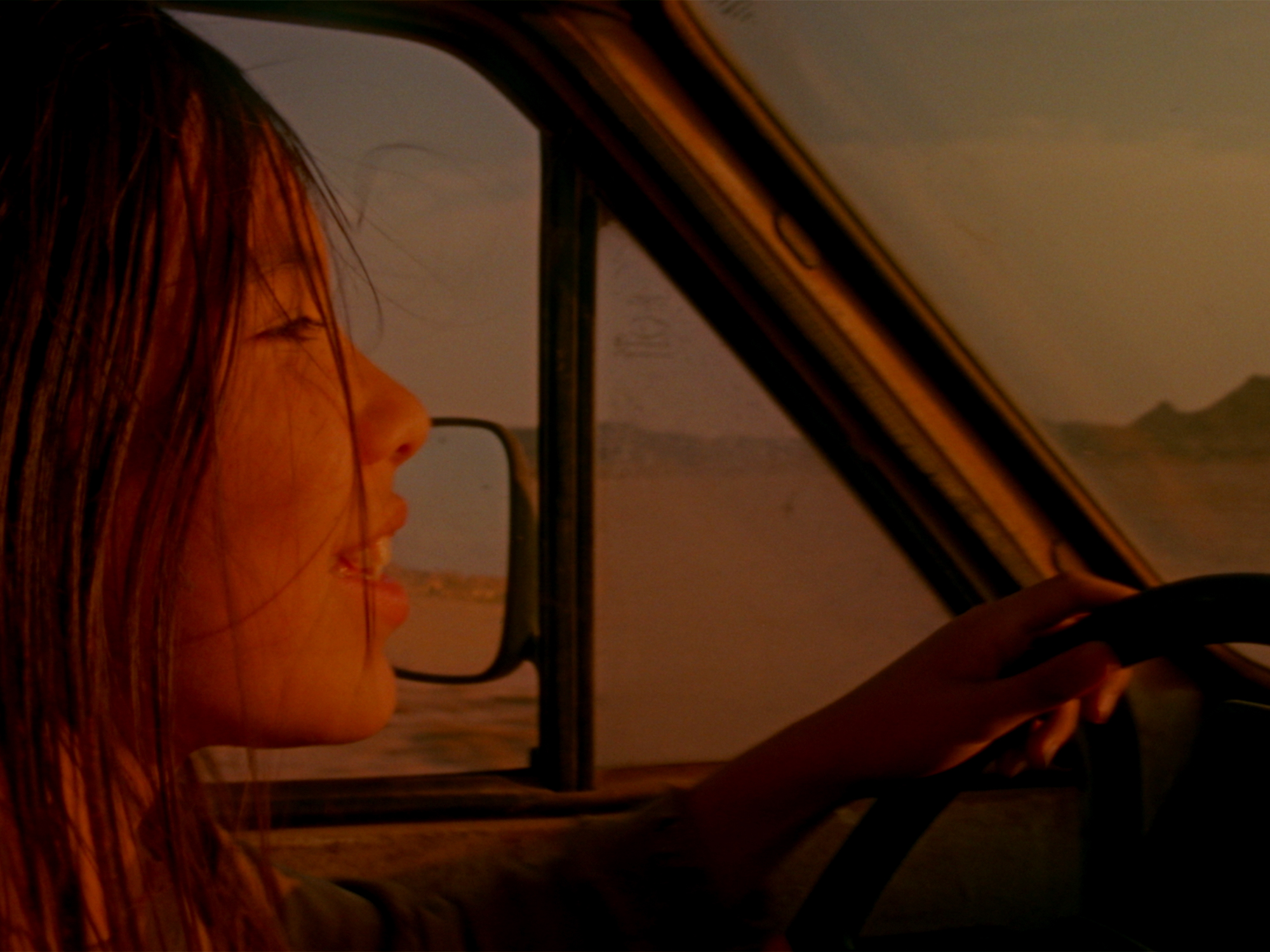 Woman with long hair driving car, viewed from passenger side. Warm orange lighting creates golden glow on face and interior.