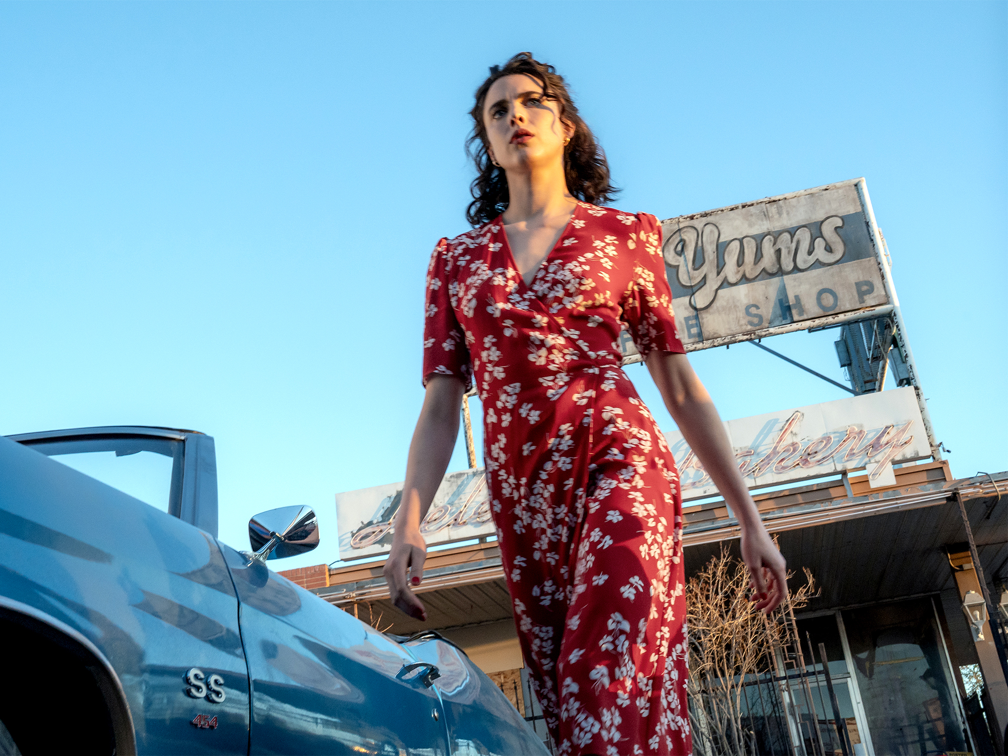 A woman in a long, red floral dress stands in front of a building with a "Gym's" sign. A car is visible in the background.