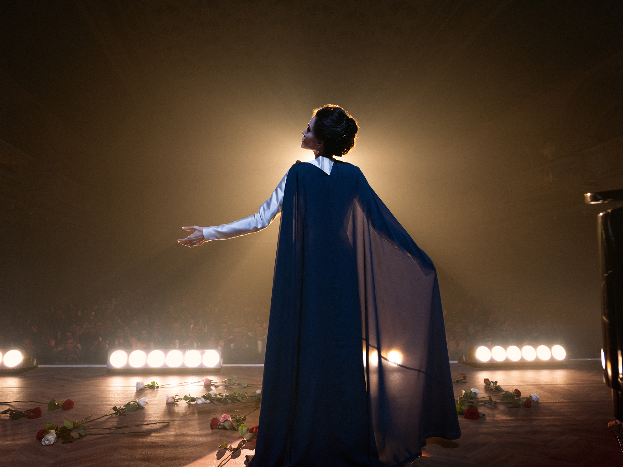 Rear view of a person wearing a dark cape standing on a stage with lights in the background.