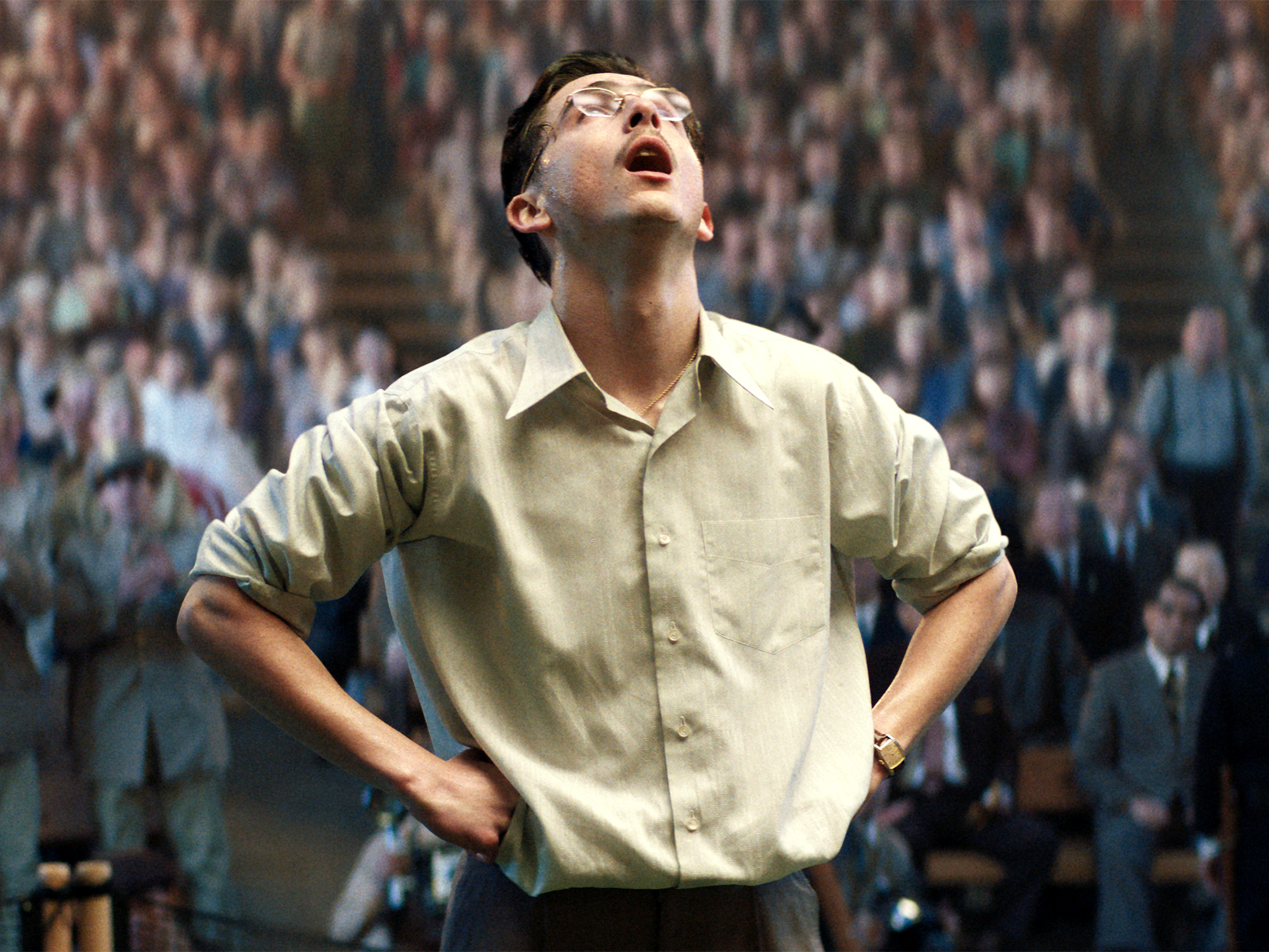 Man in cream shirt with hands on hips, head tilted back, mouth open, standing before blurred crowd in background.