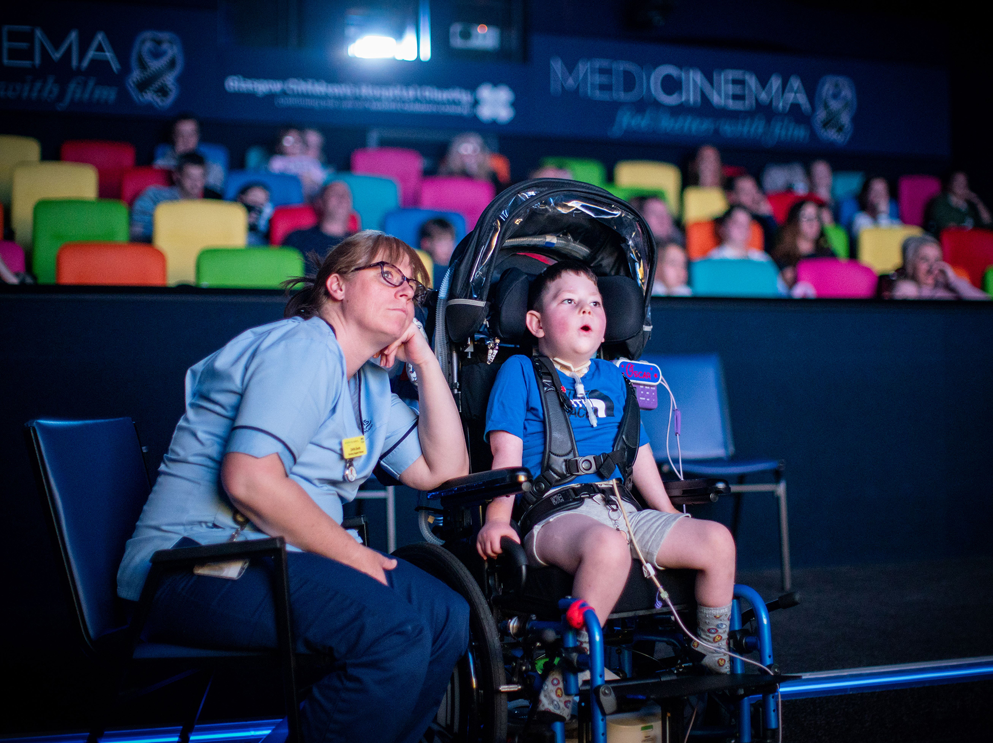 A parent and child in wheelchairs sit in a cinema auditorium with colourful seats and 'MEDICIN3MA' signage visible.