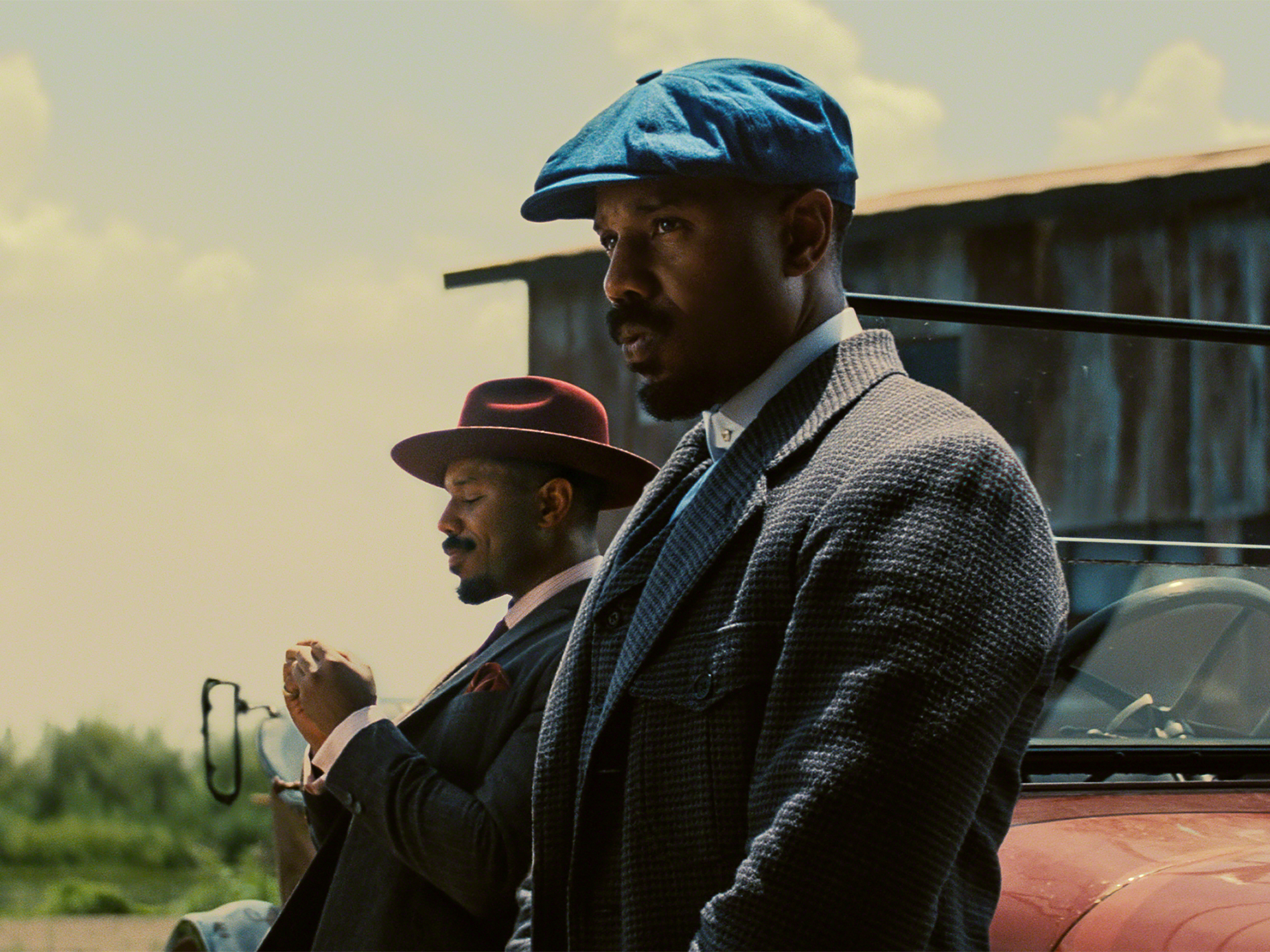Two men in period attire - one in a blue cap, the other in a cowboy hat - stand next to a vintage car in a rural setting.