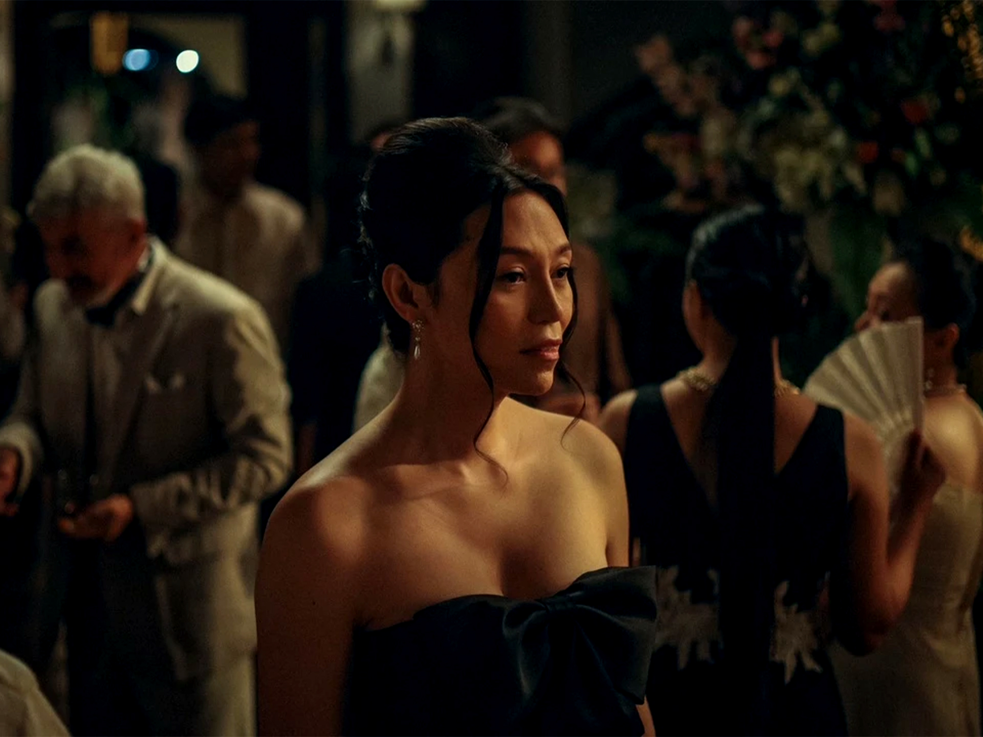 Woman in black off-shoulder dress at evening event with blurred crowd in background, warm lighting.