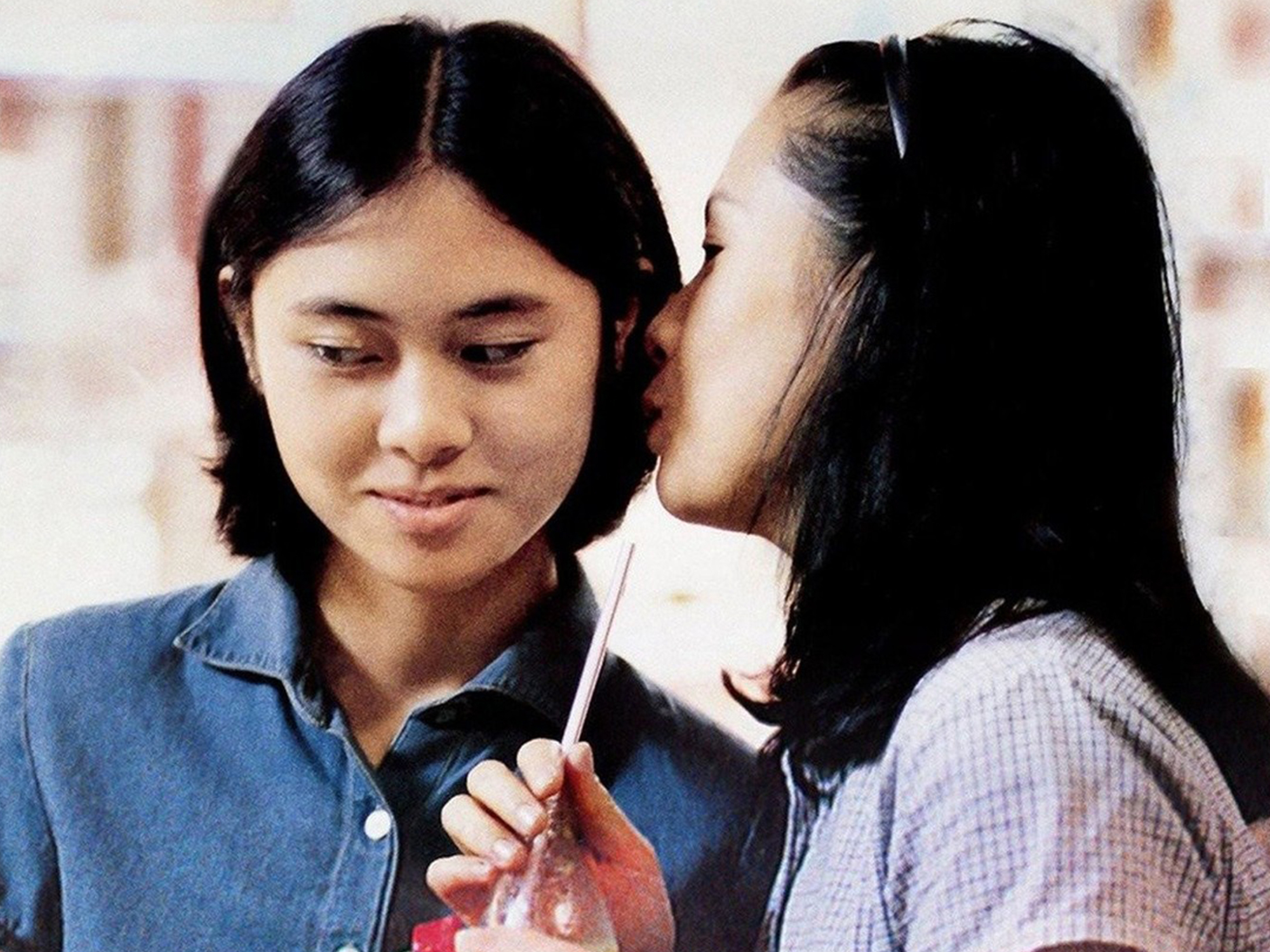 Two young Chinese women stand together, one in a blue denim shirt, the other in a purple and white gingham shirt. The one in the purple and white shirt is holding a soda bottle with a straw, and is whispering in the other girl's ear.