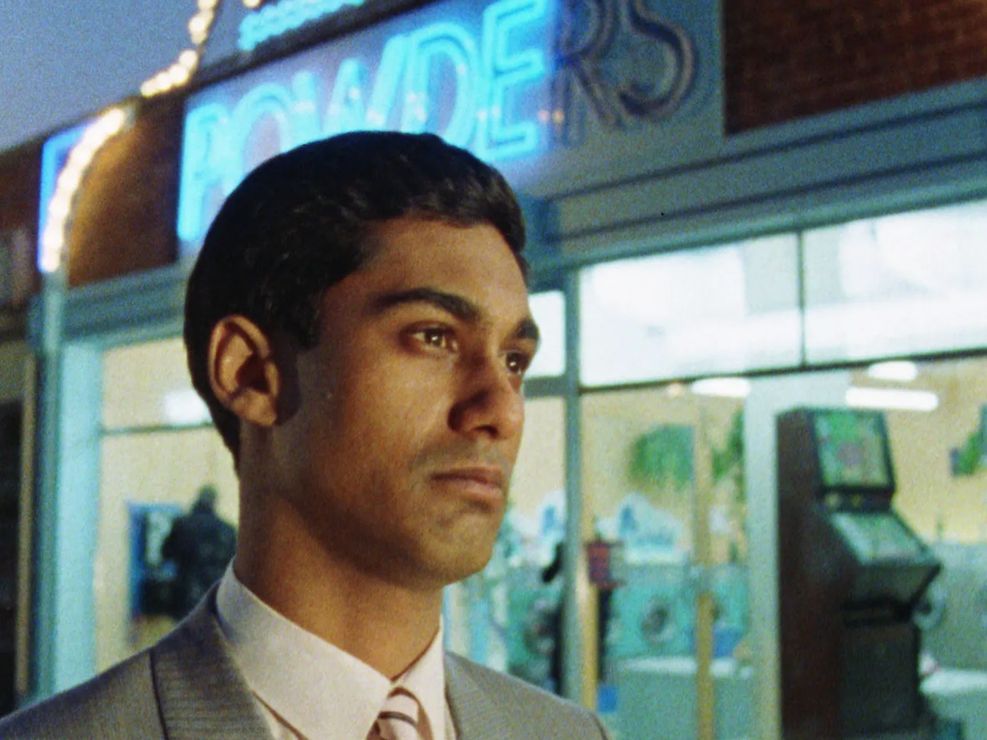 Gordon Warnecke, an Indian young man wearing a suit and tie, looking off to the side of the camera with a serious expression, against a backdrop of a launderette that has a neon sign reading "Powders"