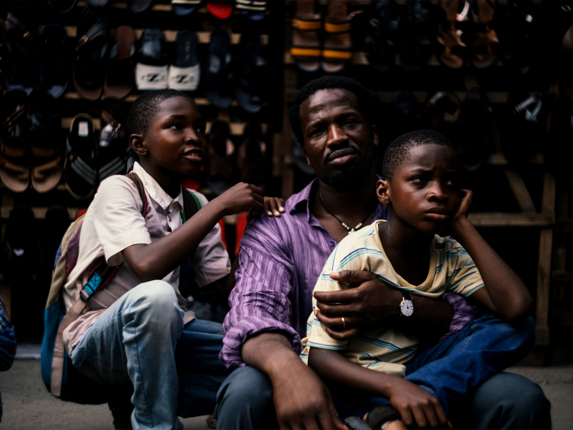 Three African men seated in a dimly lit stall, wearing casual clothing. The men appear to be in conversation, with the younger ones looking up at the older man.