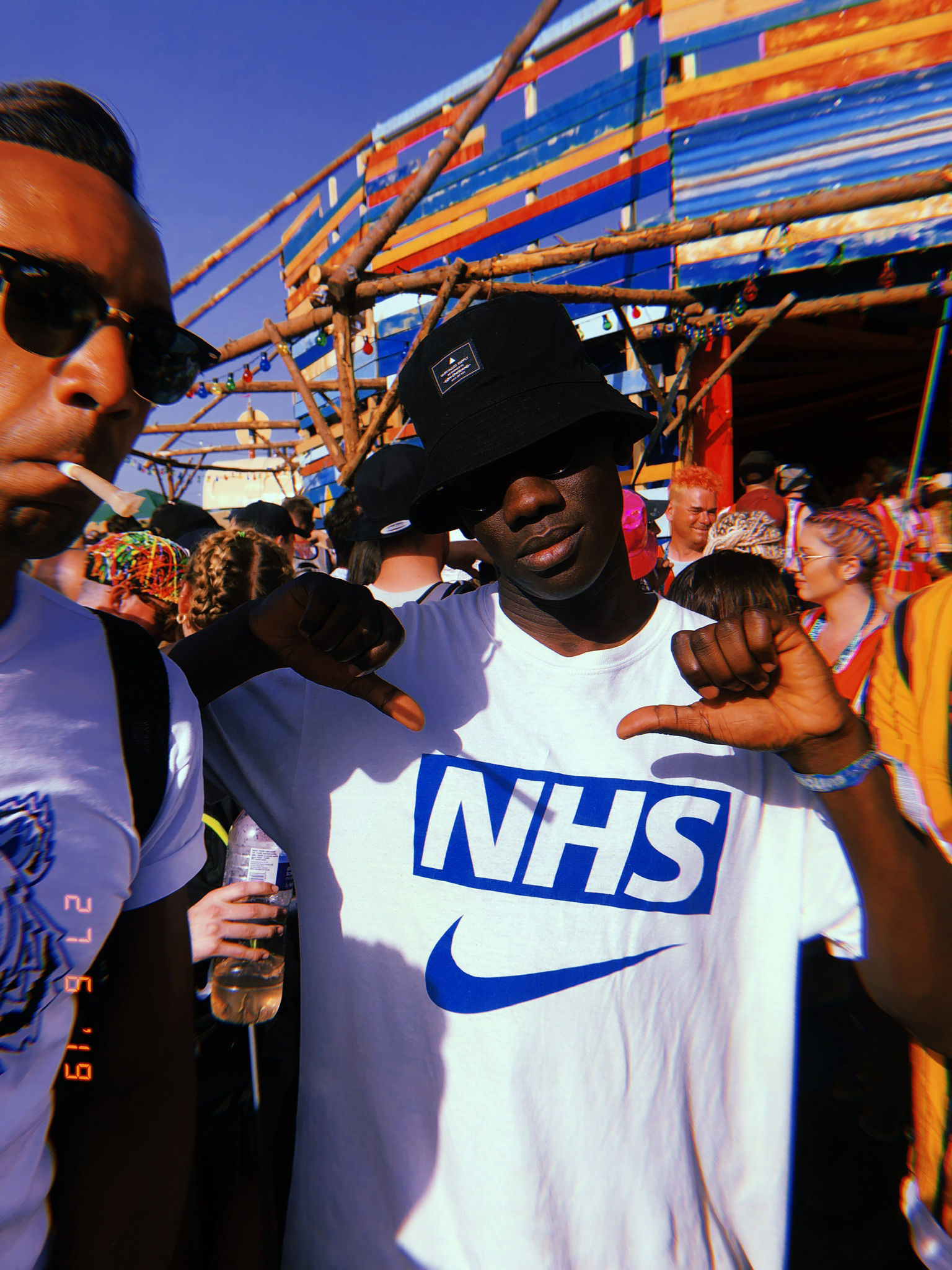 Two men at outdoor event, one wearing white NHS Nike t-shirt and black cap, colourful striped canopy overhead against blue sky.