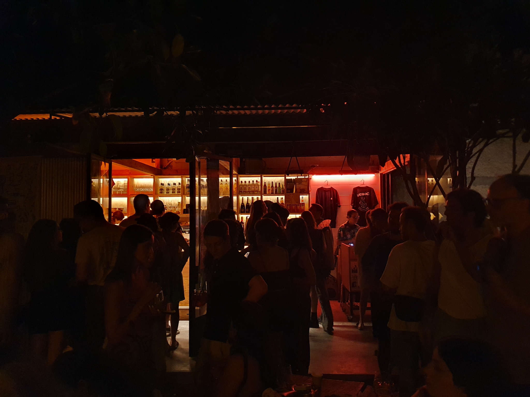 Dimly lit bar with warm orange and red lighting, silhouettes of people gathered outside and inside, bottles visible on shelves.