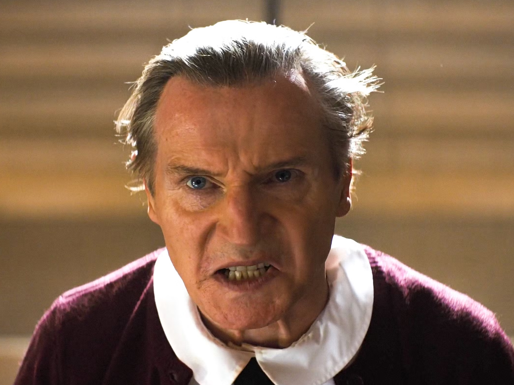 Liam Neeson in The Naked Gun. Liam Neeson is a white male in his 60s with grey hair and blue eyes. He is frowning and bearing his teeth in a grimace. He is wearing a burgundy cardigan and a round-collared white shirt.