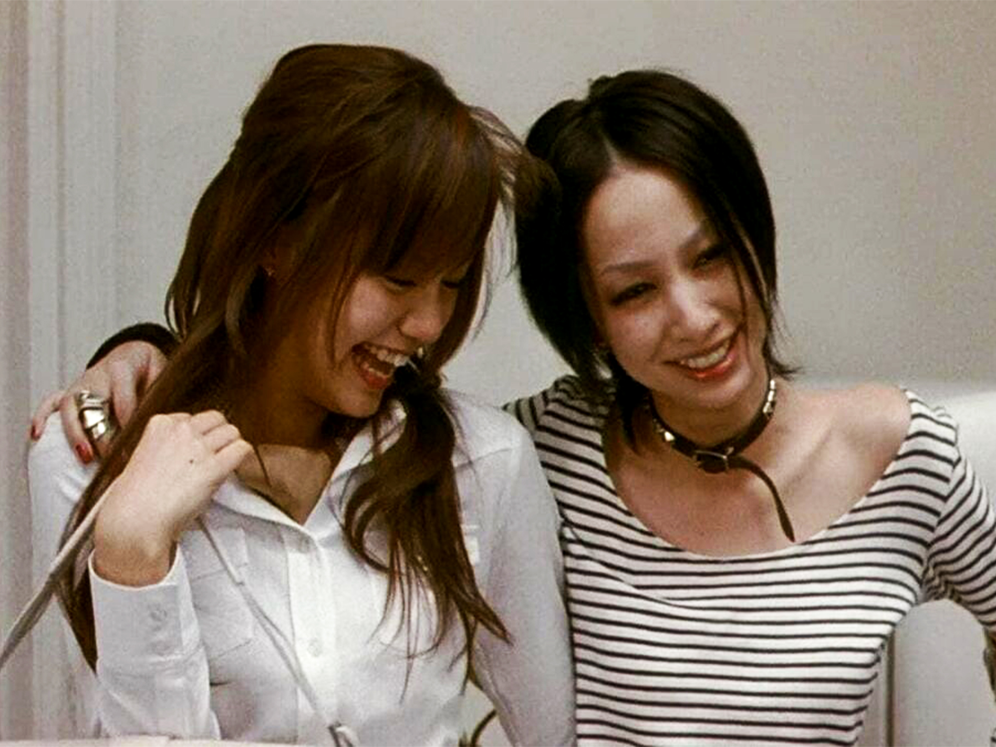 Two women laughing with arms around each other. One wears white shirt, other wears black and white striped top with necklace.