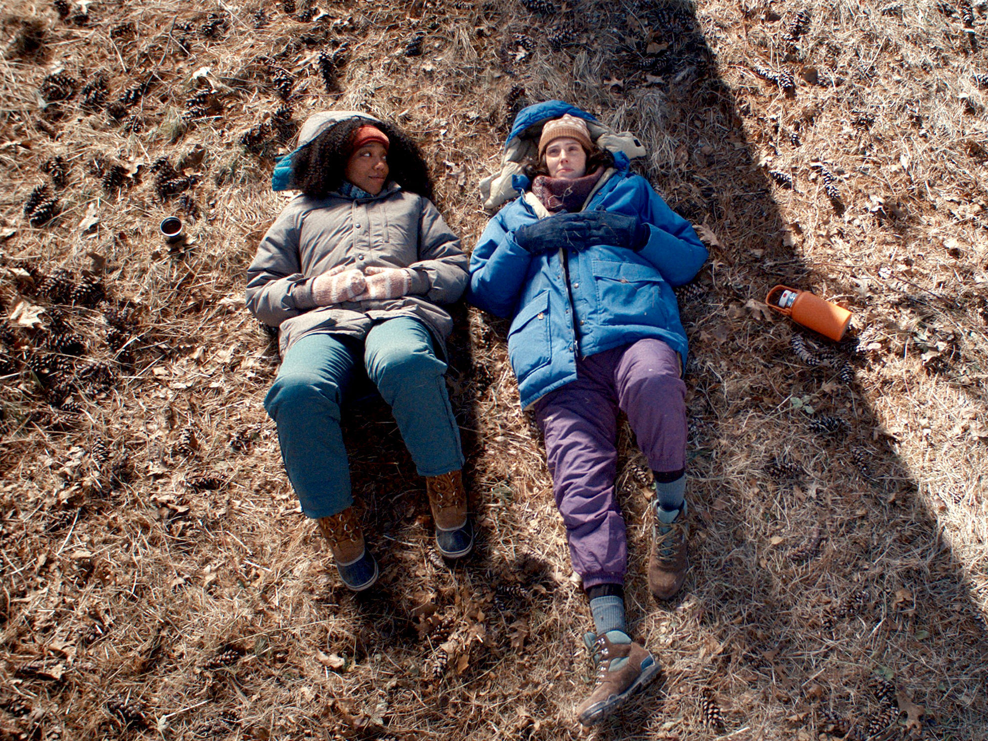 Two women lying on rocky ground in shadow, one wearing grey jumper and jeans, other in blue jacket and purple trousers.