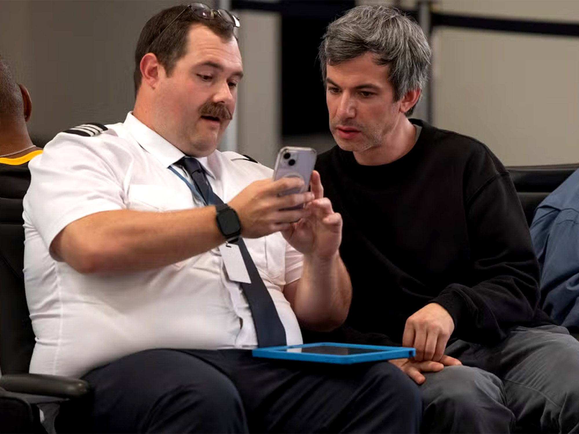 Two men seated, one in white shirt and tie holding mobile phone, other in black jumper looking at screen together.
