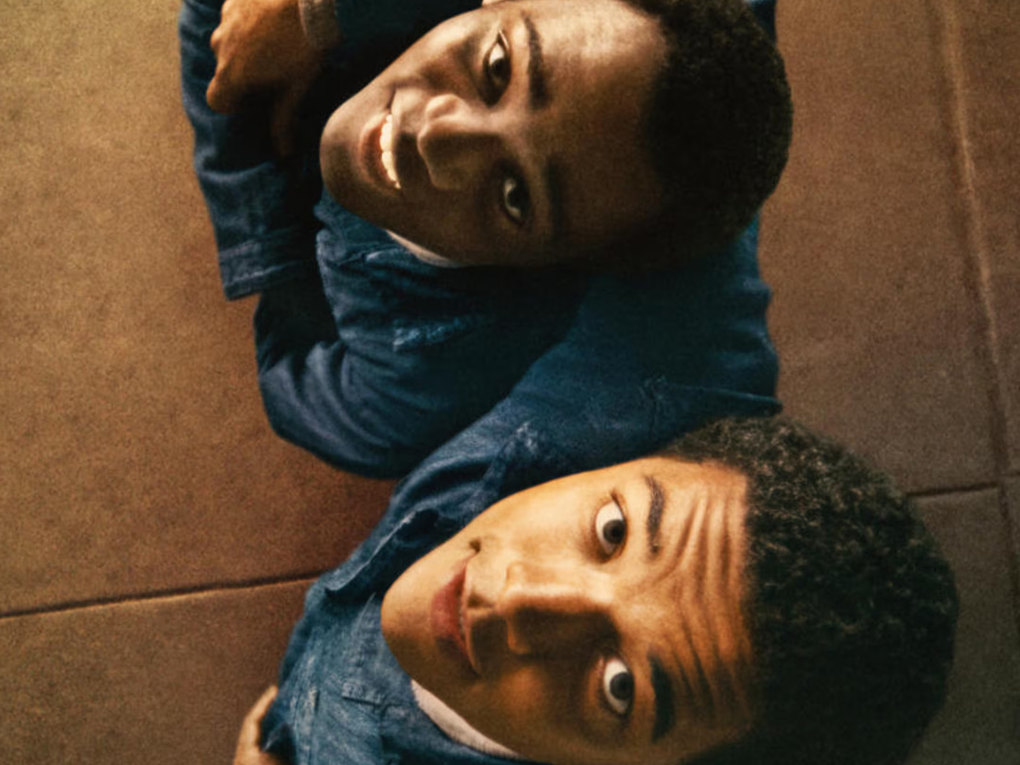 A close-up image of two young people, a man and a woman, lying on the floor and looking at the camera with neutral expressions.