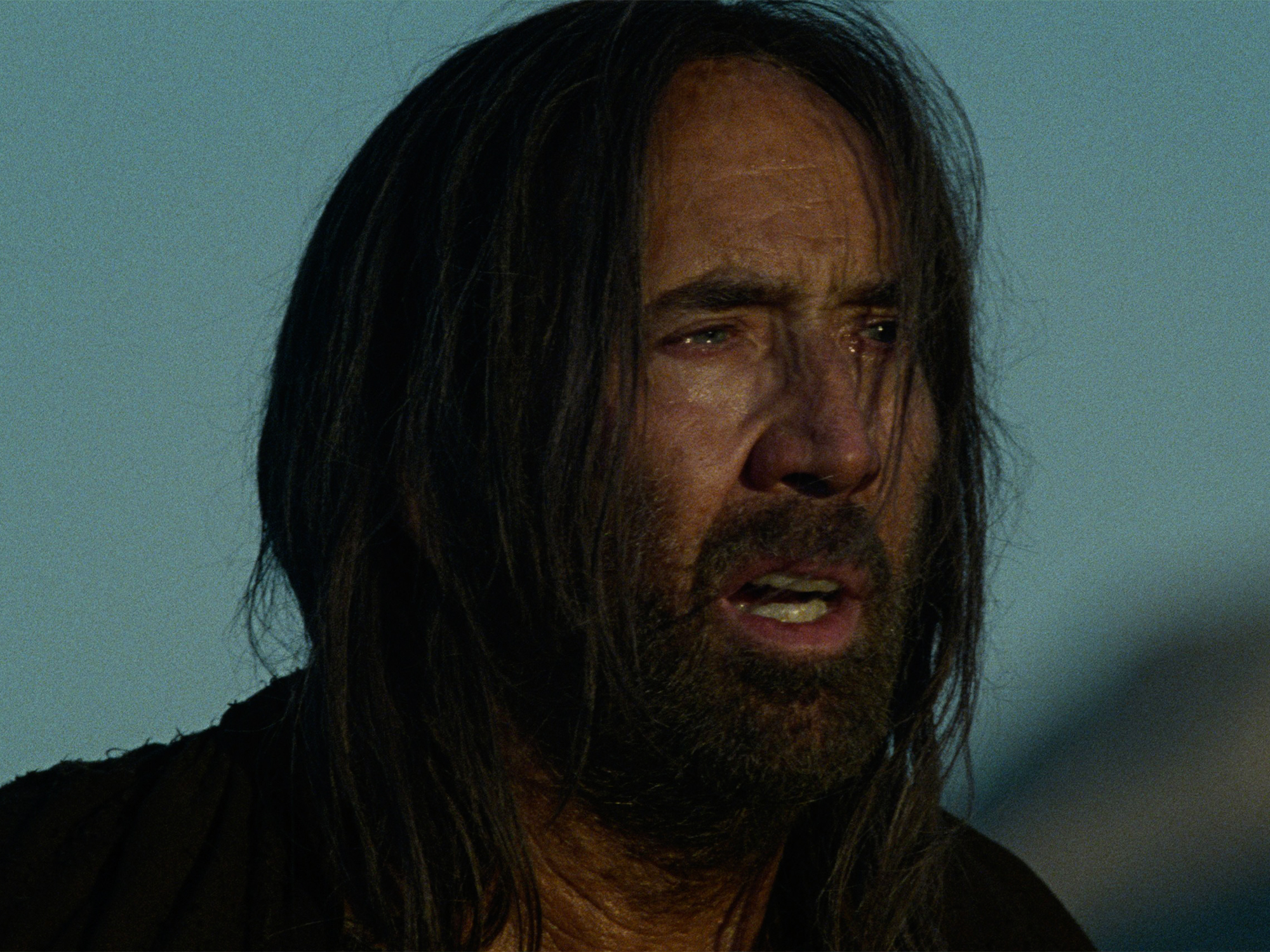 Man with long dark hair and beard against blue-grey sky background, mouth slightly open, wearing dark clothing.