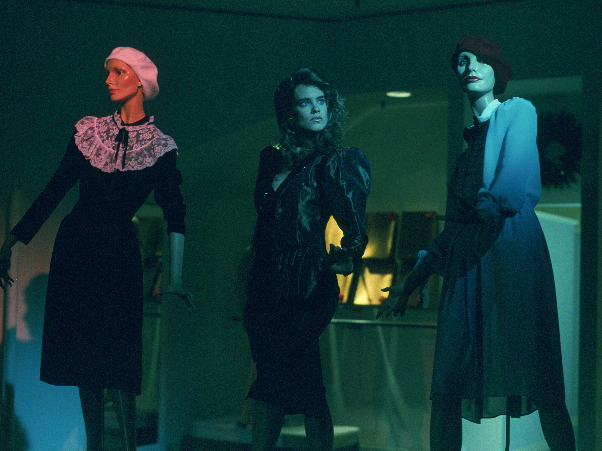 Three women in period costumes stand in green-tinted lighting. Left wears black dress with pink ruffled collar, centre in dark gown, right in blue top and dark skirt.