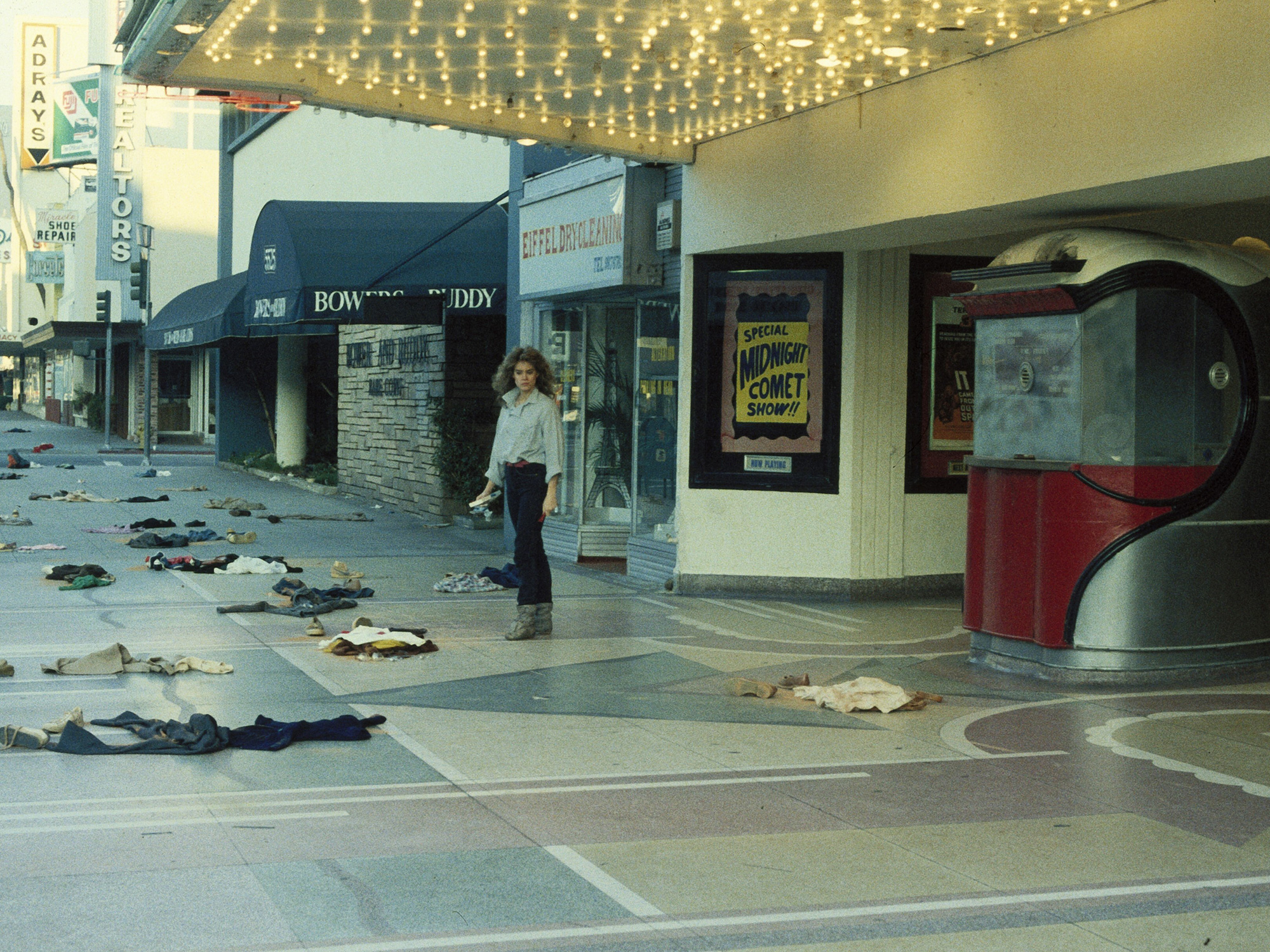 Shopping centre with debris scattered across tiled floor, person walking past storefronts, overhead lighting visible, some shops appear damaged.
